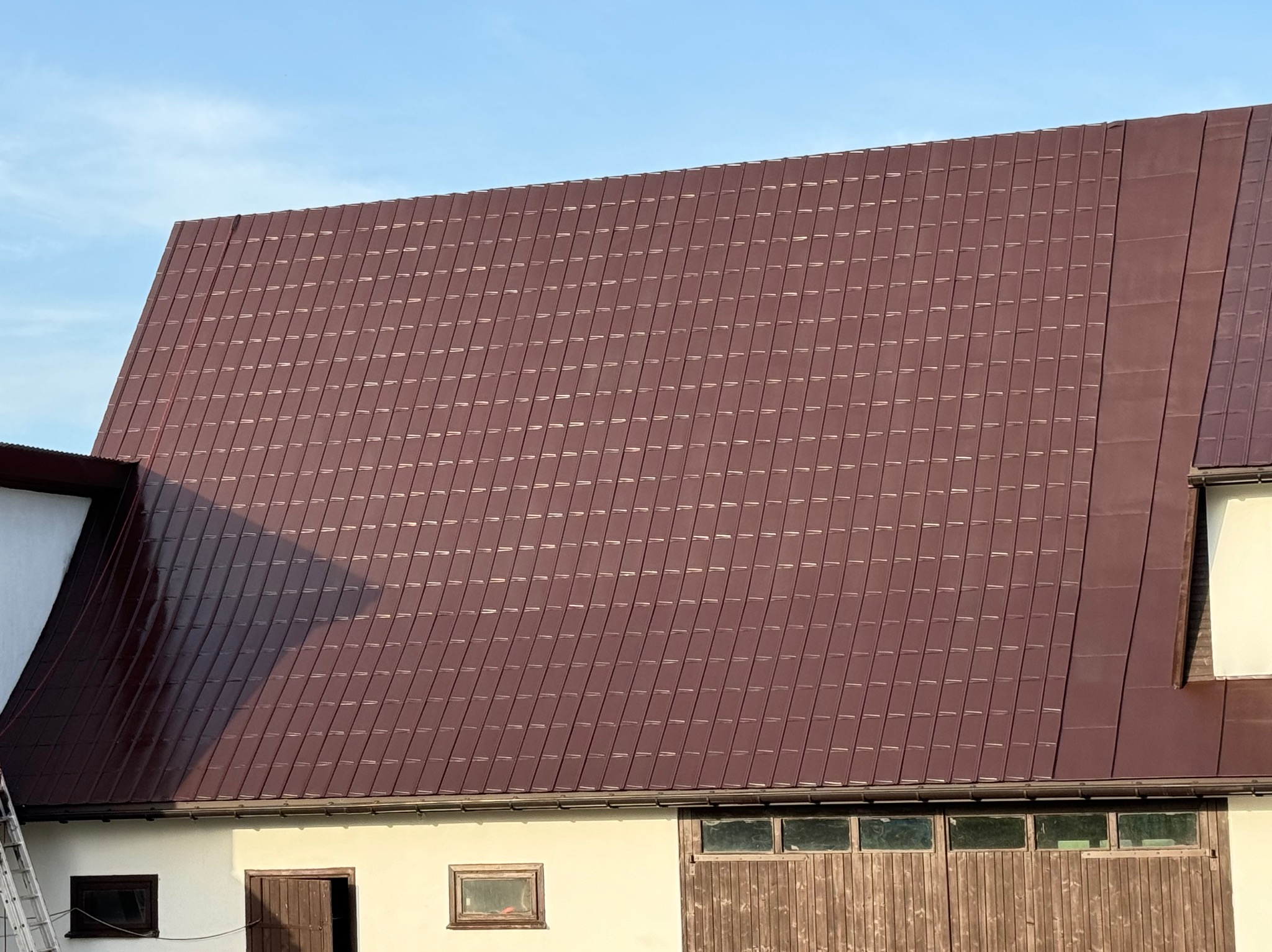 Ciemnobrązowy dach z blachy trapezowej na budynku gospodarczym, widoczne okna i drewniana brama, fragment drabiny po lewej stronie.
