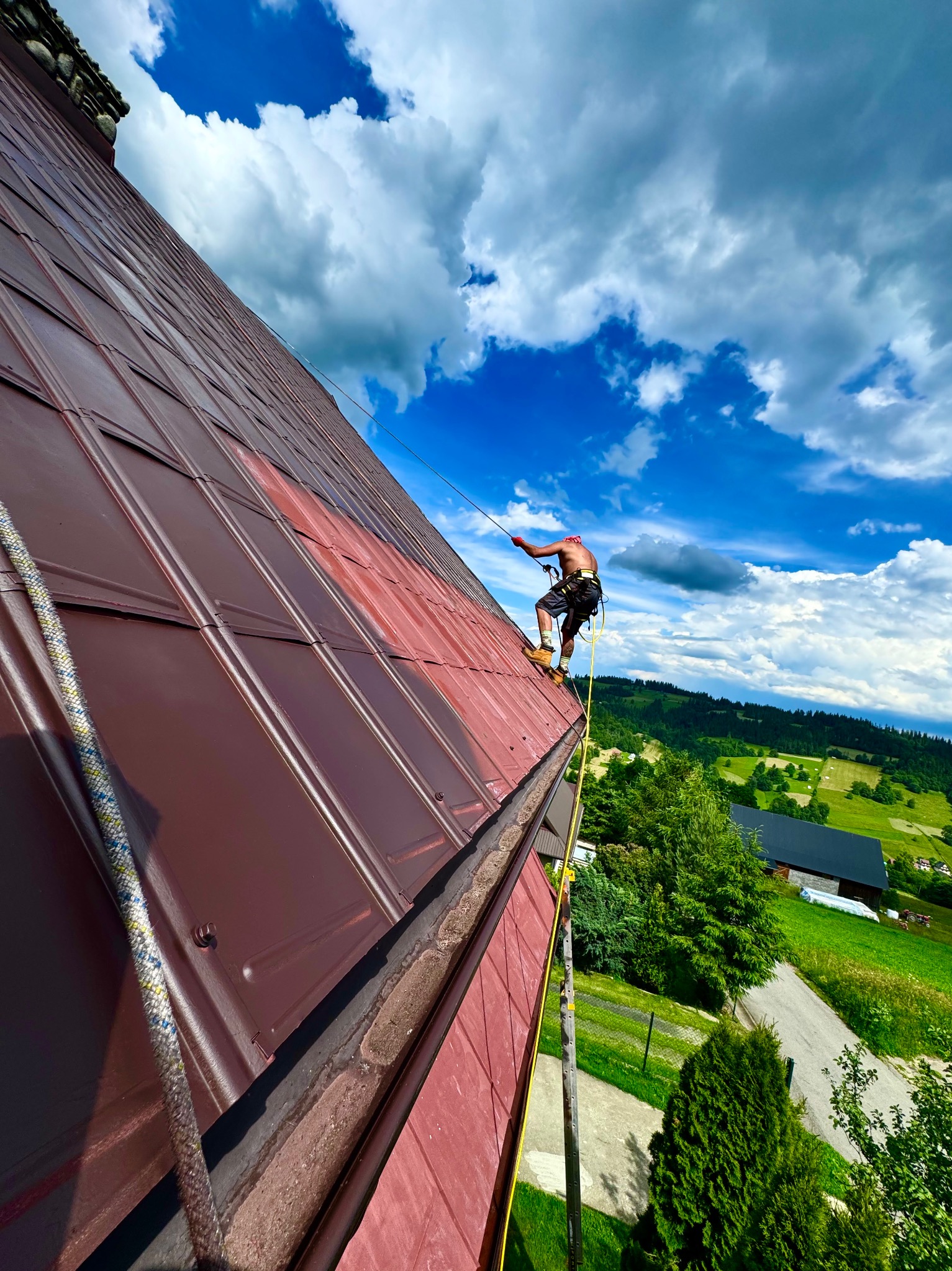 Pracownik na stromym dachu z blachy na tle błękitnego nieba z chmurami, zabezpieczony linami, częściowo pomalowany dach w kolorze ceglastym, widok na zielone wzgórza w tle.