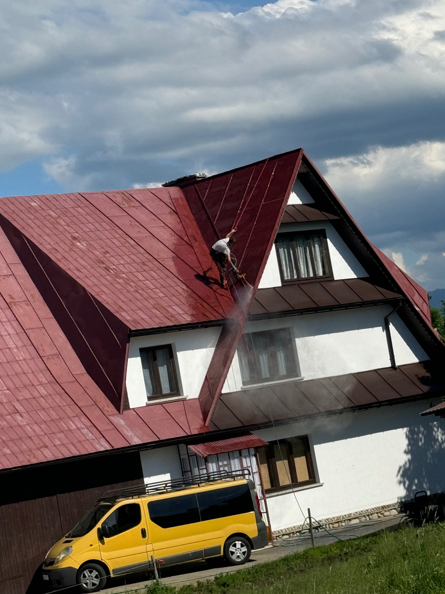 Pracownik zabezpieczony linami, czyści dach domu z czerwonej dachówki za pomocą myjki ciśnieniowej, widoczny żółty bus firmy na tle białej elewacji.