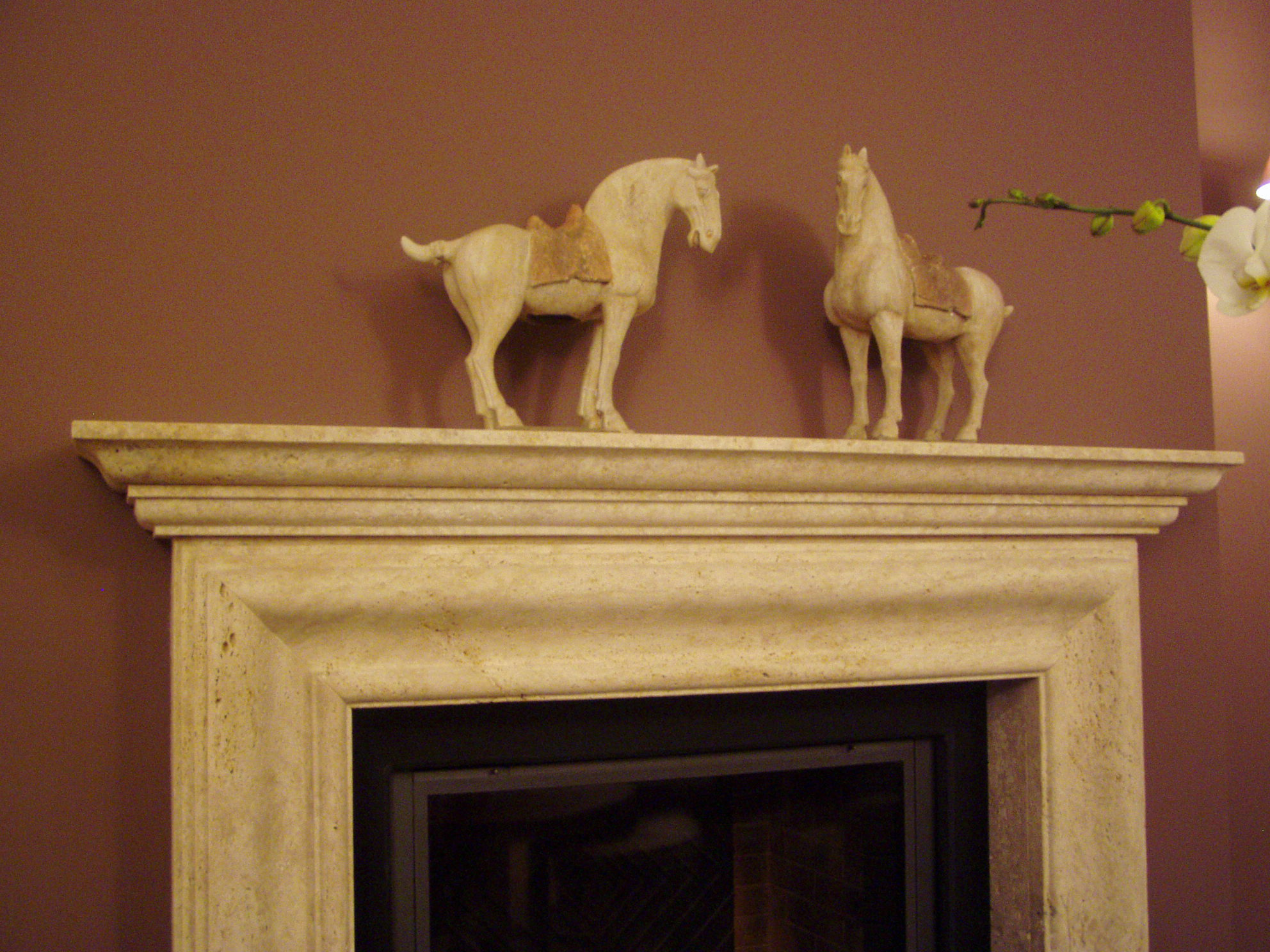 Obudowa kominka z jasnego kamienia z dekoracyjnymi figurkami koni i gałązką orchidei na górnej półce.