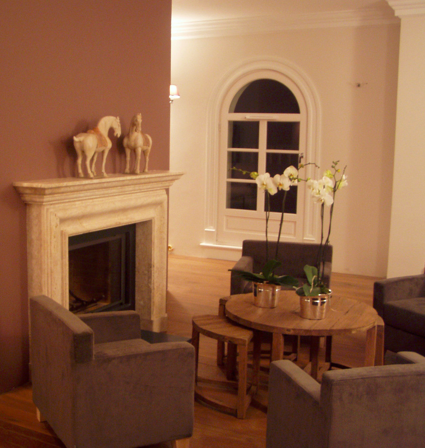 Salon z marmurowym kominkiem, figurkami koni na gzymsie, drewnianym stolikiem z orchideami w srebrnych doniczkach i szarymi fotelami na drewnianej podłodze.