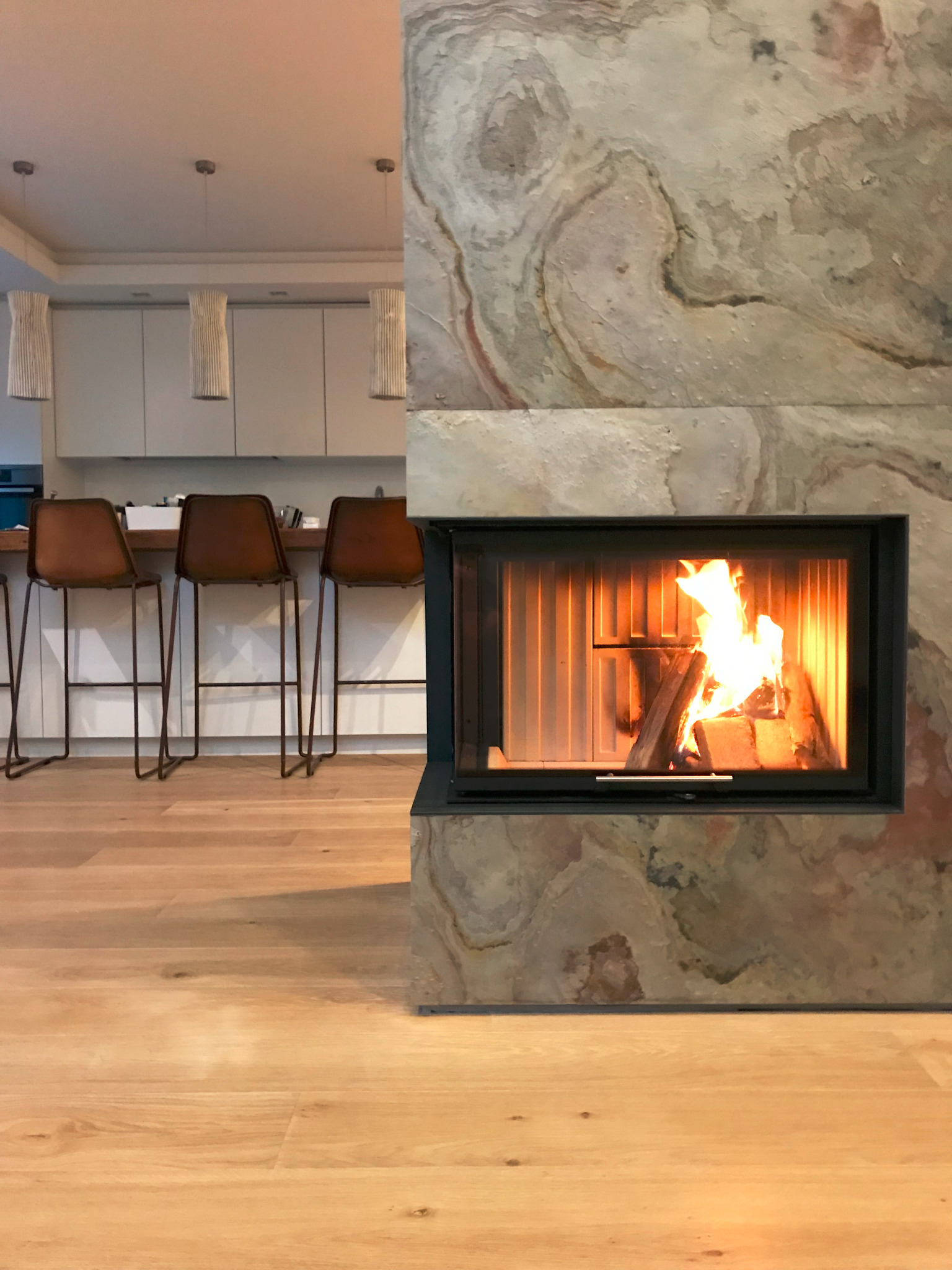 Nowoczesny kominek obudowany kamieniem z paleniskiem i płomieniami, widok na kuchnię z hokerami i drewnianą podłogą.