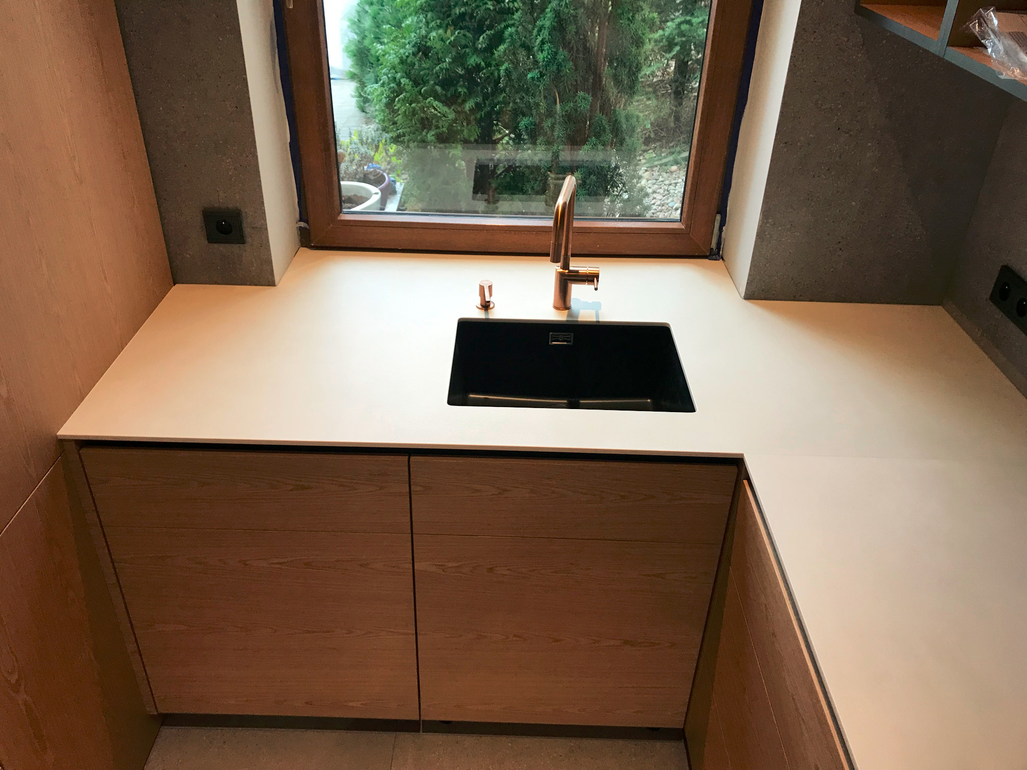 Jasny blat kuchenny z czarnym zlewozmywakiem i miedzianą baterią, widok na ogród przez okno.