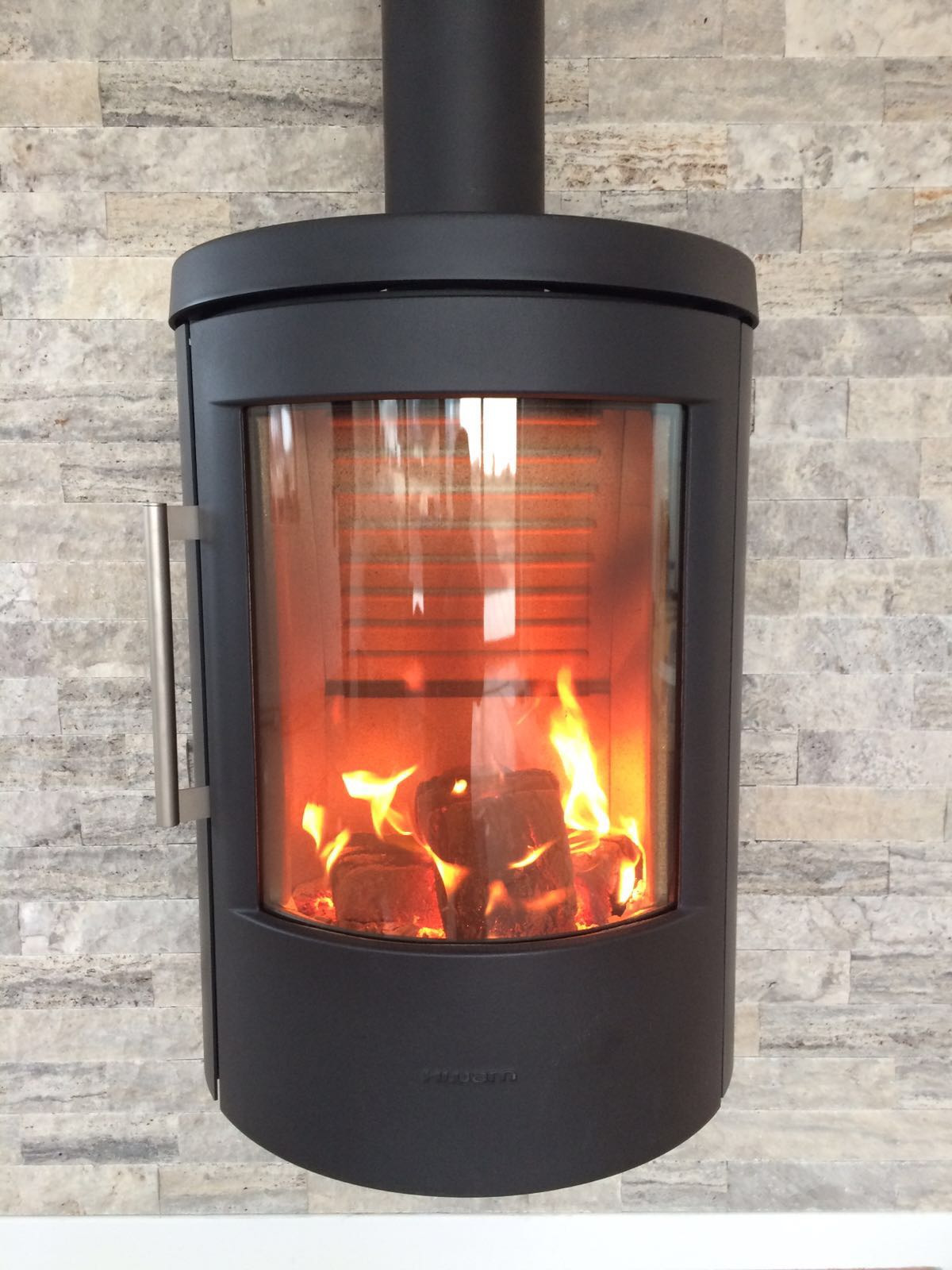 Nowoczesny, wiszący, czarny piec kominkowy z płomieniami, zamontowany na ścianie wyłożonej dekoracyjnym kamieniem w odcieniach szarości.