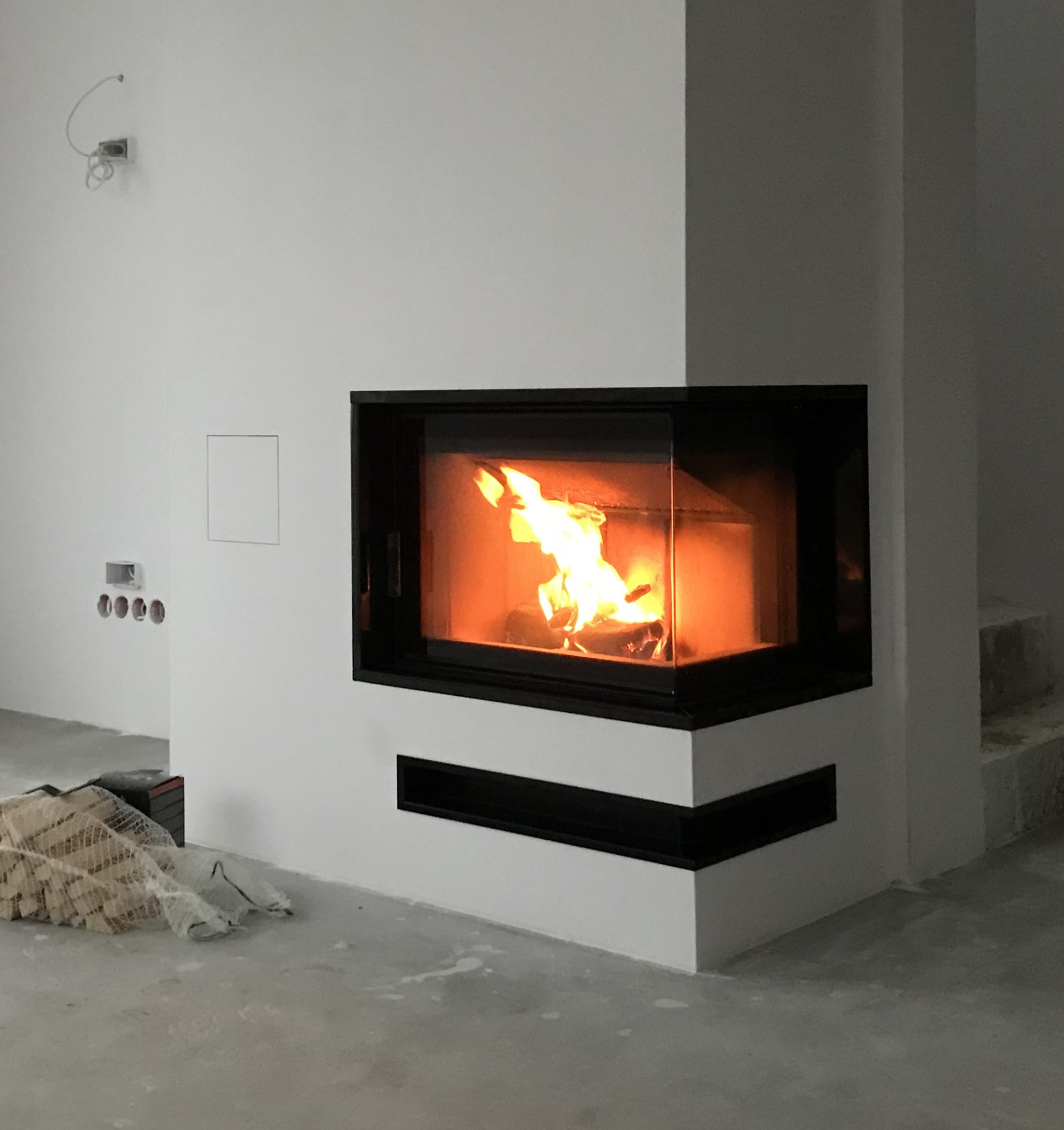 Nowoczesny kominek narożny z czarną obudową i paleniskiem z płomieniami, widoczne przygotowane drewno obok, surowe wykończenie podłogi i ściany w tle.