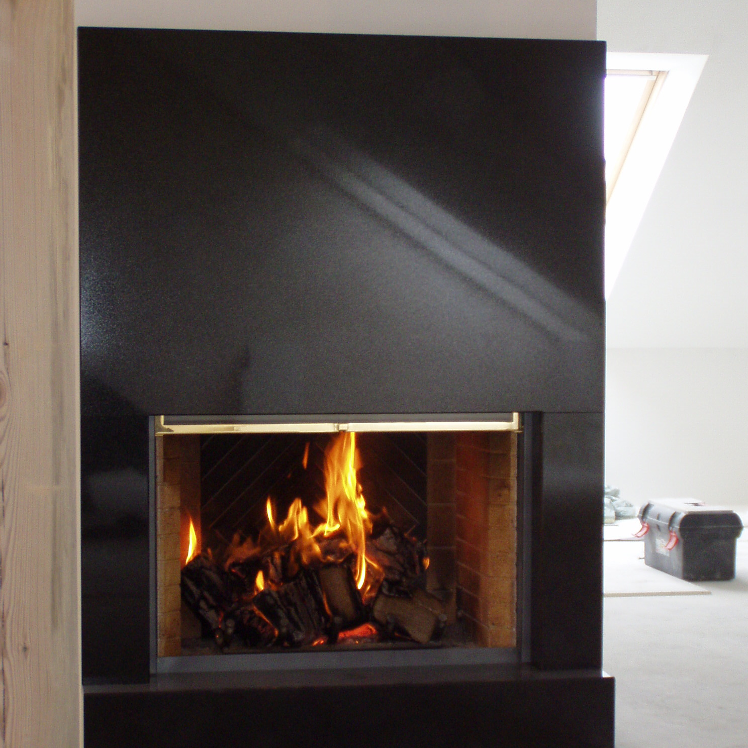 Czarny, nowoczesny kominek z paleniskiem wypełnionym płonącym drewnem, widoczne narzędzia i fragment okna dachowego w tle.