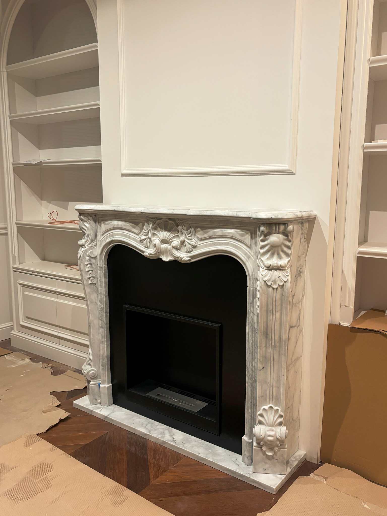 Marmurowy kominek z dekoracyjnymi rzeźbieniami w trakcie montażu, widoczne zabezpieczenia podłogi i fragmenty regałów w tle.