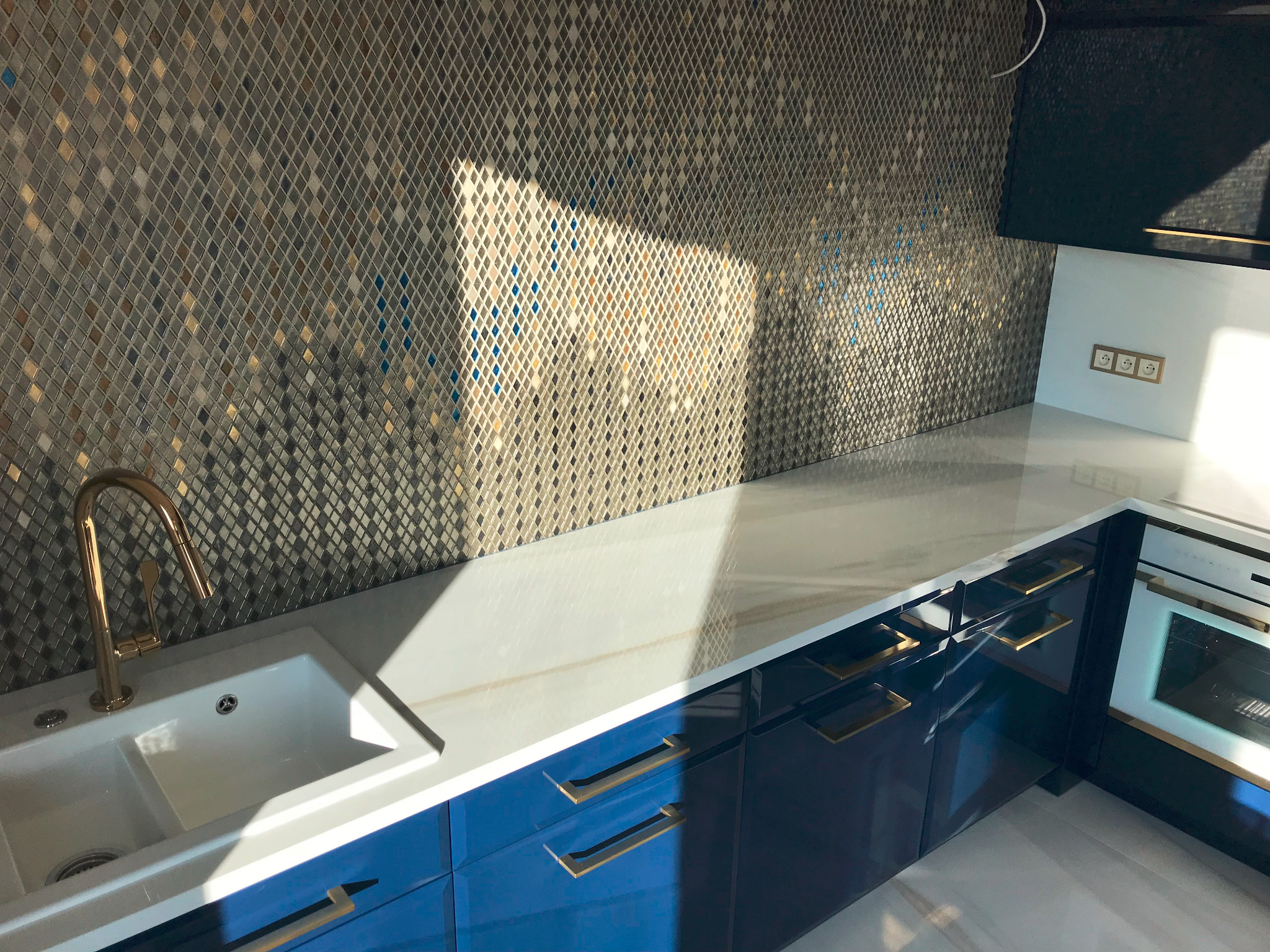 Błyszcząca kuchnia z mozaikową ścianą, białym blatem i niebieskimi szafkami z złotymi uchwytami, oświetlona promieniami słońca.