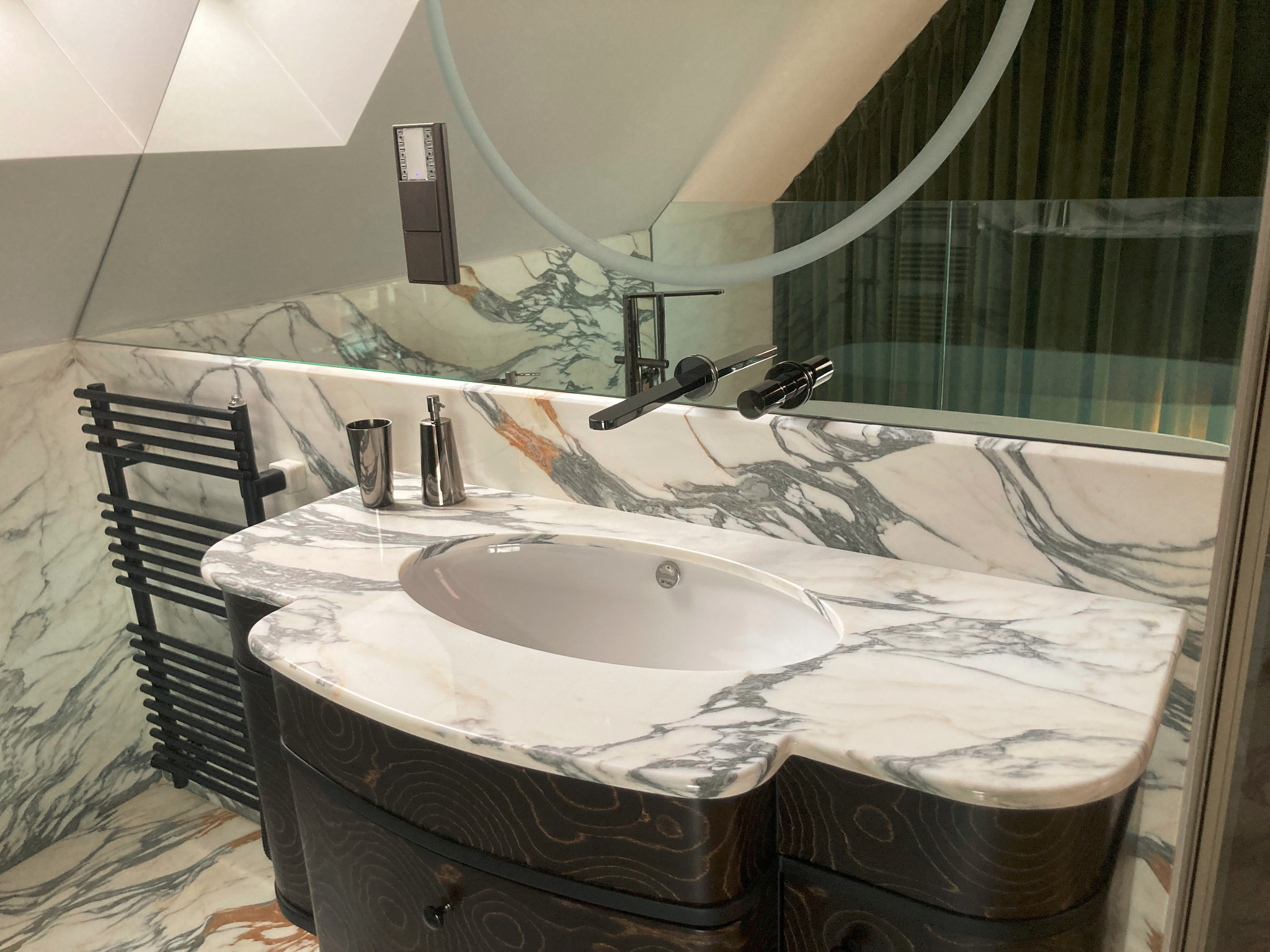 Nowoczesna łazienka z marmurowym blatem, owalną umywalką i czarną armaturą, uzupełniona grzejnikiem drabinkowym i akcesoriami w metalicznych odcieniach.