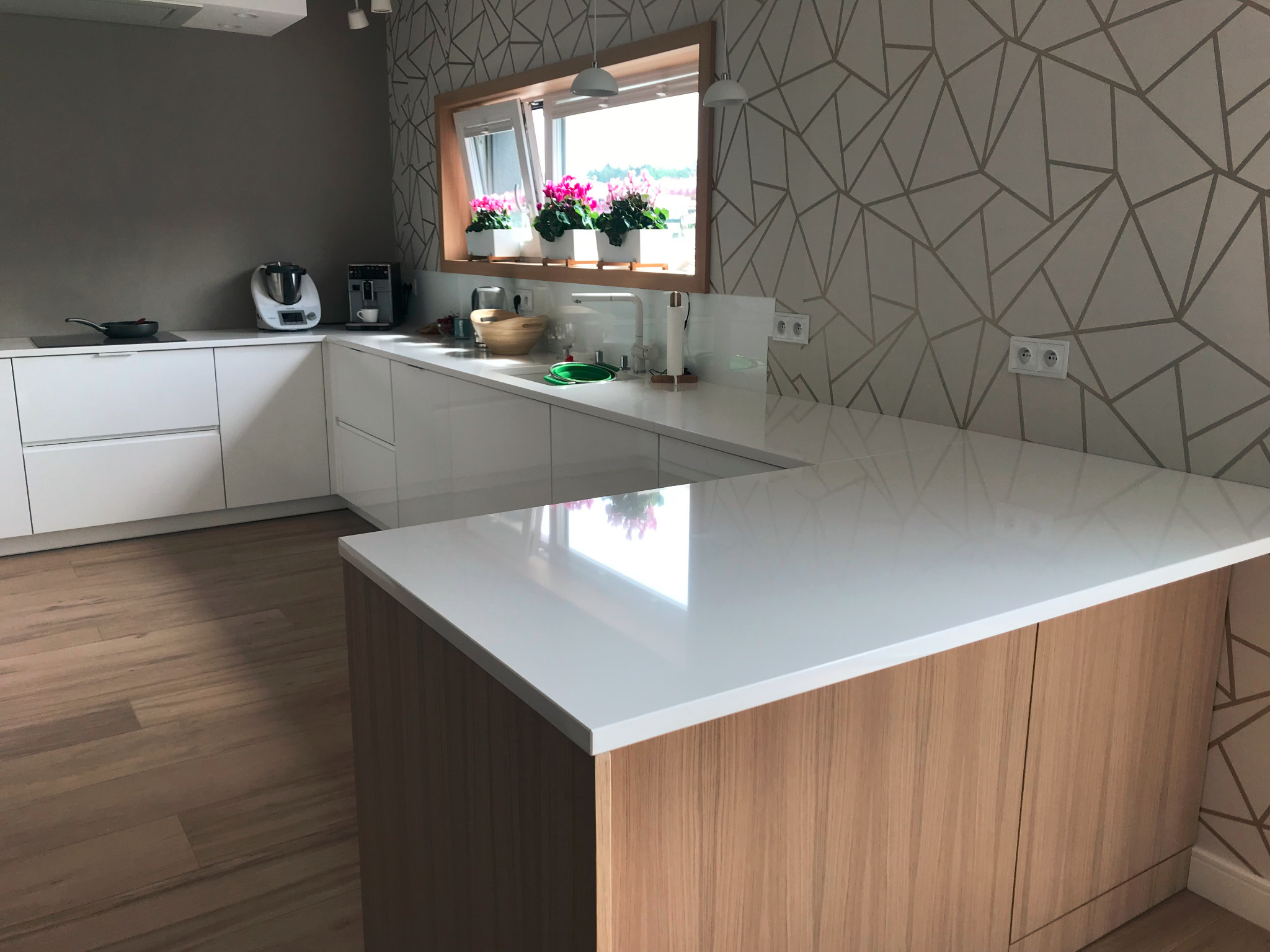 Nowoczesna kuchnia z białymi szafkami, jasnym drewnianym blatem wyspy i geometryczną tapetą w tle, oświetlona światłem dziennym wpadającym przez okno z kwiatami.