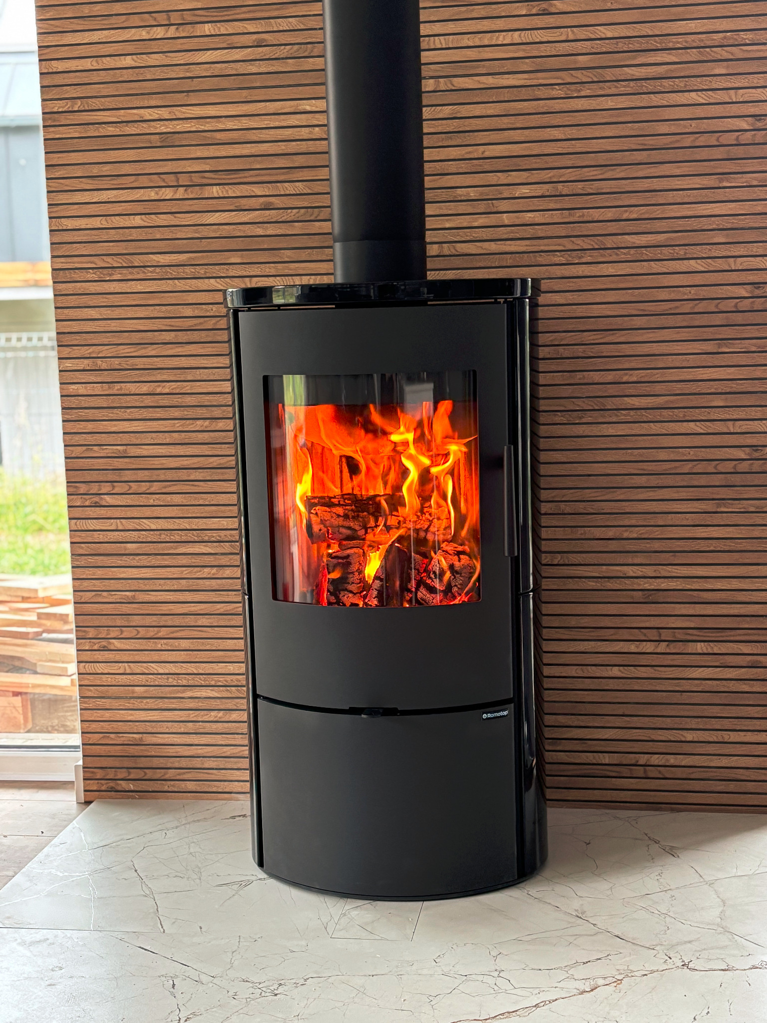 Czarny, nowoczesny kominek wolnostojący Romotop z płomieniami, na tle drewnianej ściany i marmurowej podłogi. Widoczne palące się drewno wewnątrz.