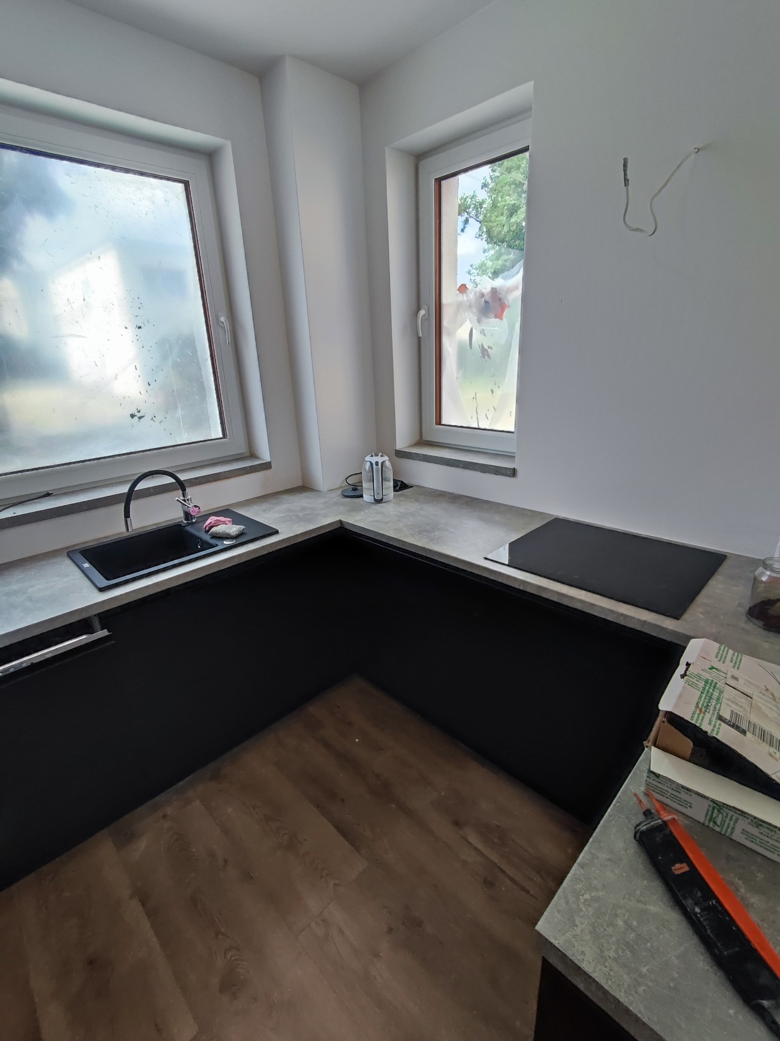 Częściowo ukończona, narożna kuchnia z czarnymi szafkami, szarym blatem, płytą indukcyjną i czarnym zlewem pod oknem, widoczne narzędzia i niedokończone wykończenie ścian.