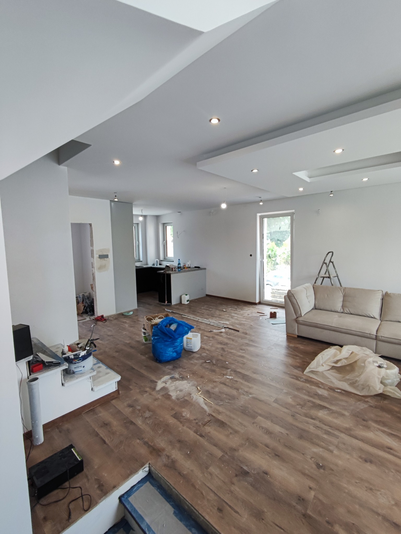 Przestronne wnętrze w trakcie prac wykończeniowych: świeżo położone panele podłogowe w odcieniach brązu, białe ściany i sufit z wbudowanym oświetleniem, widoczny fragment kuchni i salonu z beżową...