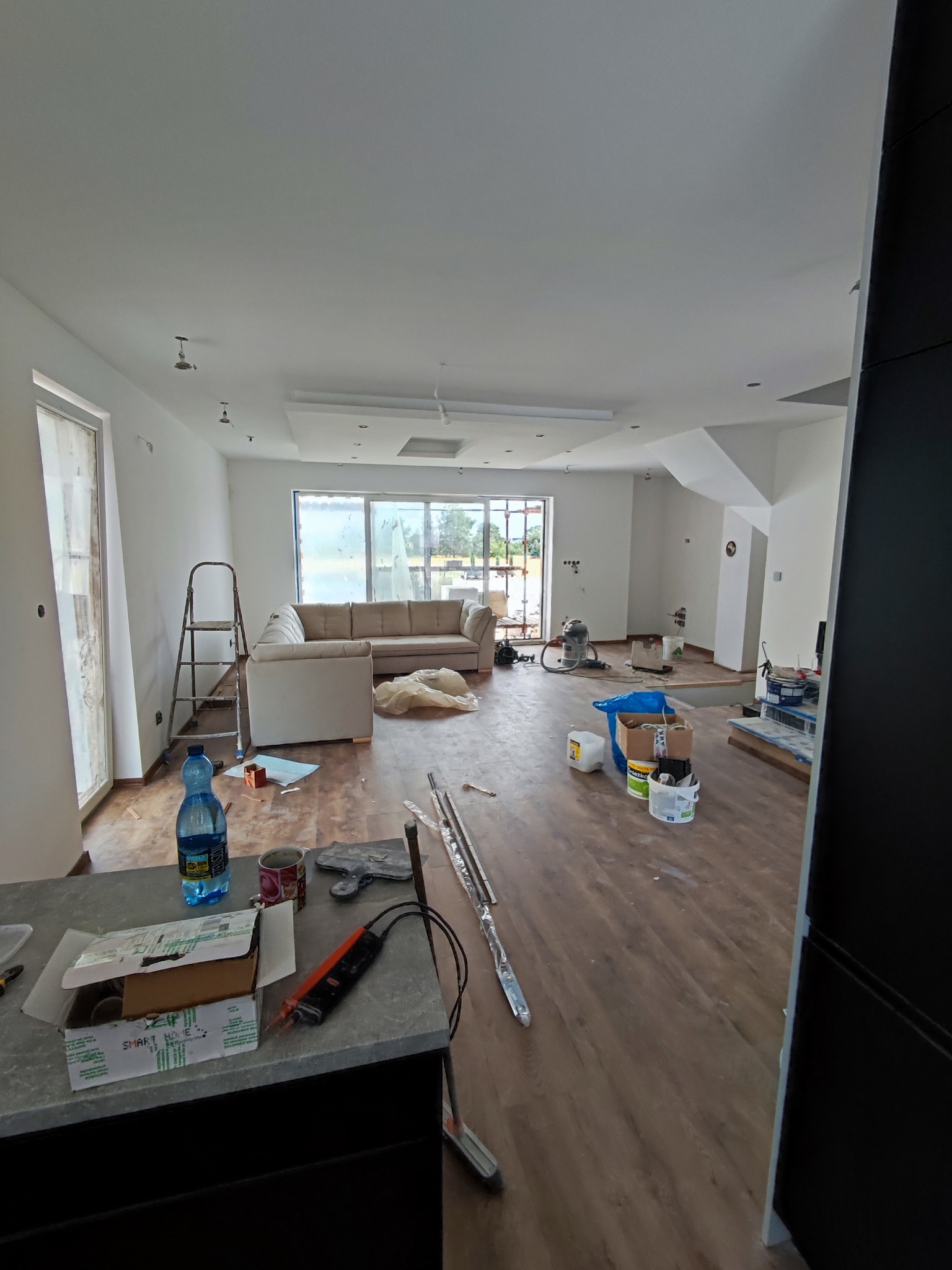 Wnętrze w trakcie prac wykończeniowych: widoczny salon z położonymi panelami podłogowymi, przygotowany do malowania, z częściowo zakrytymi meblami, narzędziami i materiałami budowlanymi.