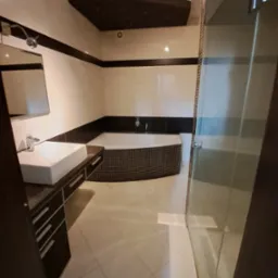 Nowoczesna łazienka z wanną obudowaną mozaiką, białą umywalką nablatową i szklaną kabiną prysznicową, utrzymana w jasnej kolorystyce z czarnymi akcentami.