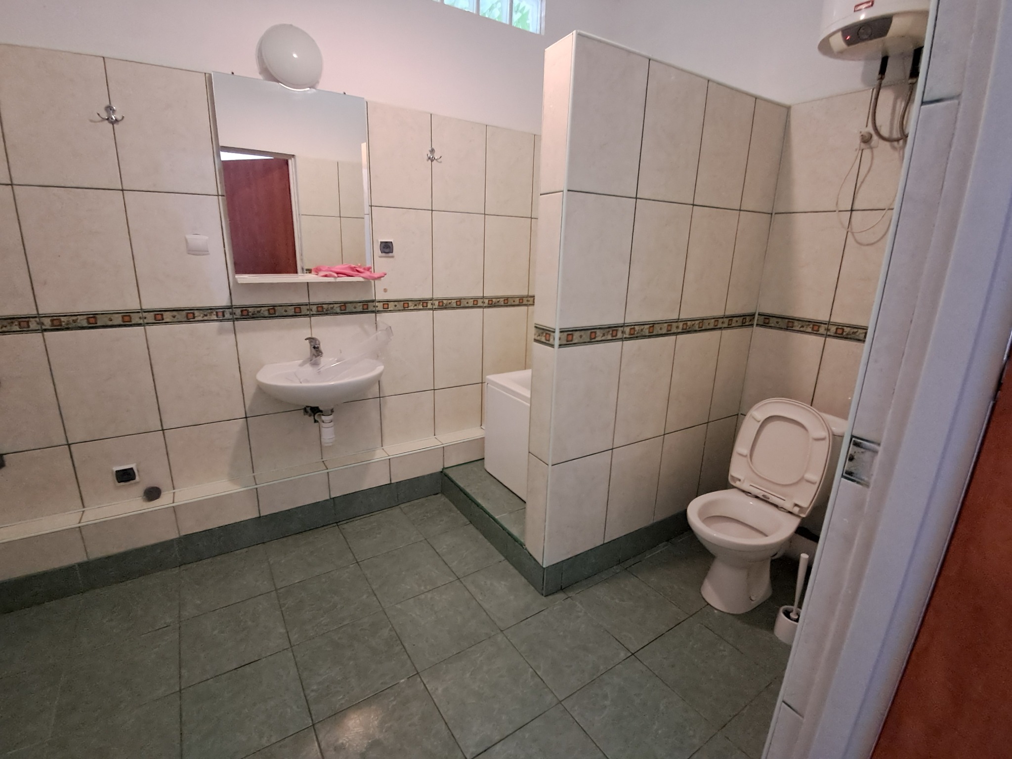 Wnętrze łazienki z beżowymi płytkami, umywalką, toaletą i bojlerem, widok z perspektywy wejścia.