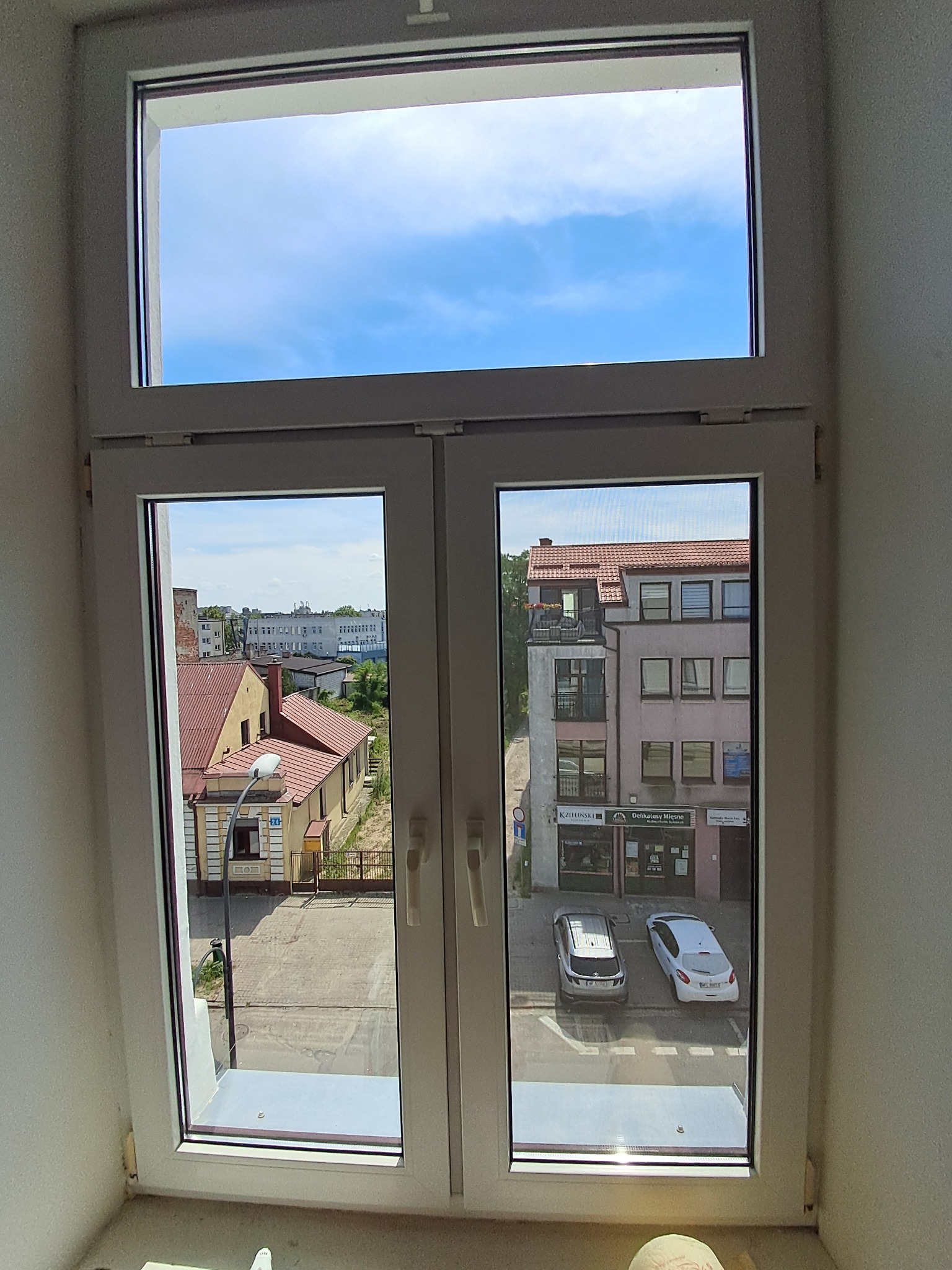 Dwuskrzydłowe, białe okno PCV z widokiem na fragment miasta: budynki mieszkalne z czerwonymi dachami, zaparkowane samochody oraz błękitne niebo.