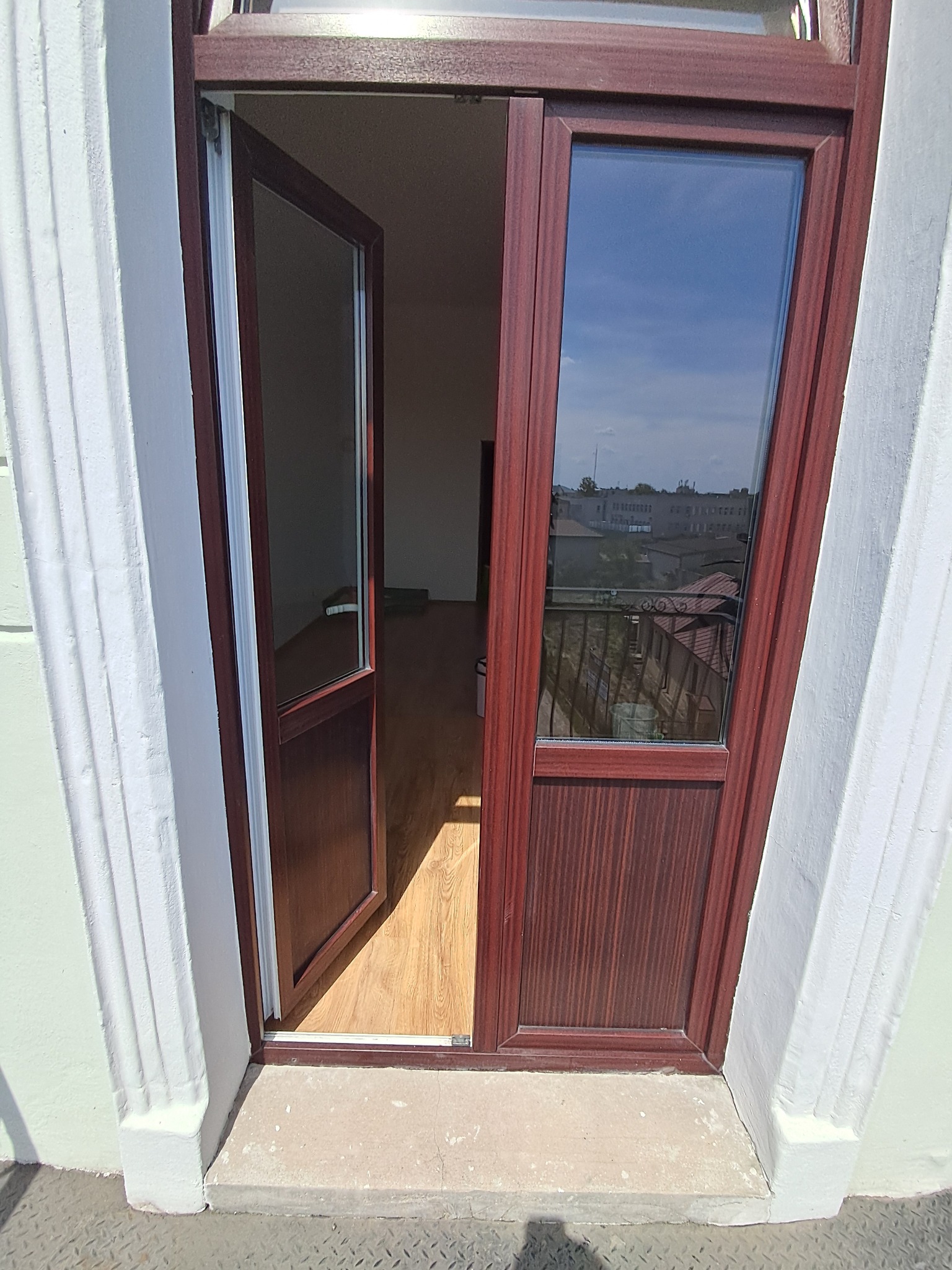 Otwarta, brązowa rama okienna z widokiem na balkon i miasto w tle, jasne panele podłogowe wewnątrz.