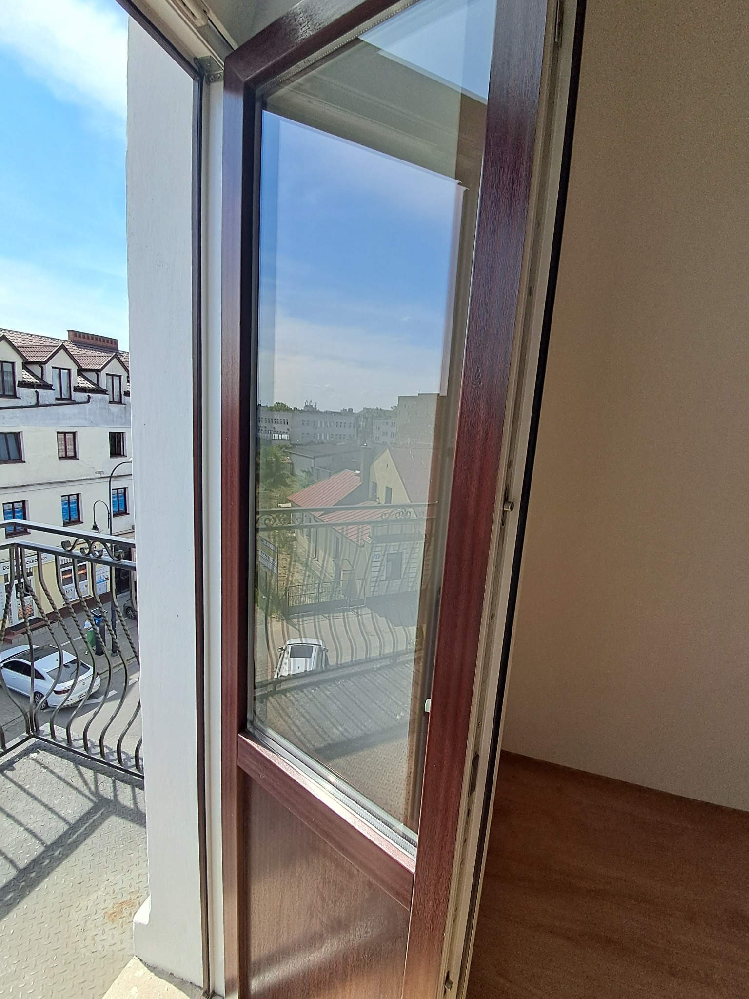 Otwarty balkon z widokiem na ulicę i budynki, czysta szyba okienna odbija otoczenie.