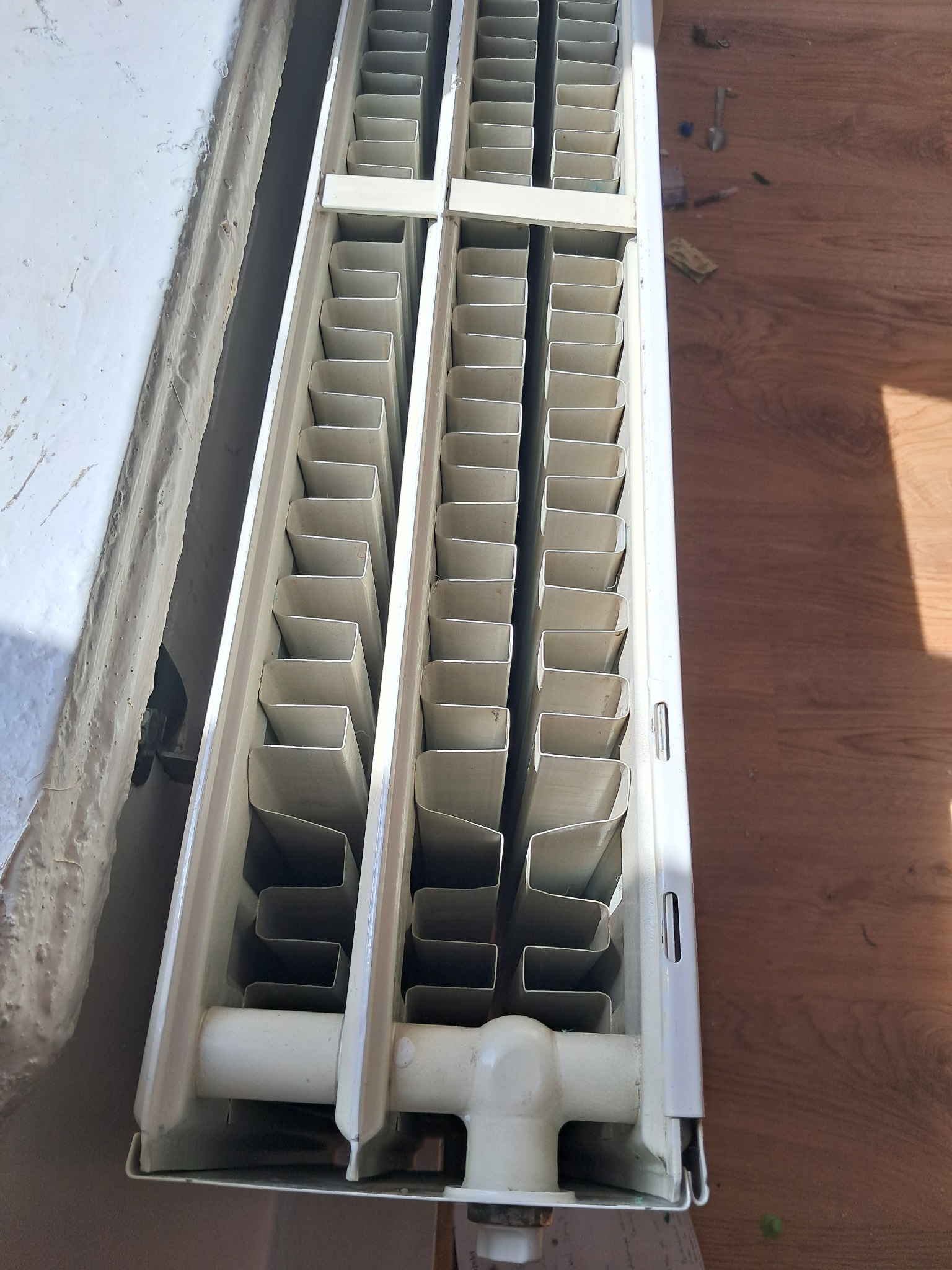 Biały, żeberkowy grzejnik zamontowany przy brudnej ścianie, widoczne ślady starej farby i zabrudzenia, na podłodze leżą drobne narzędzia.