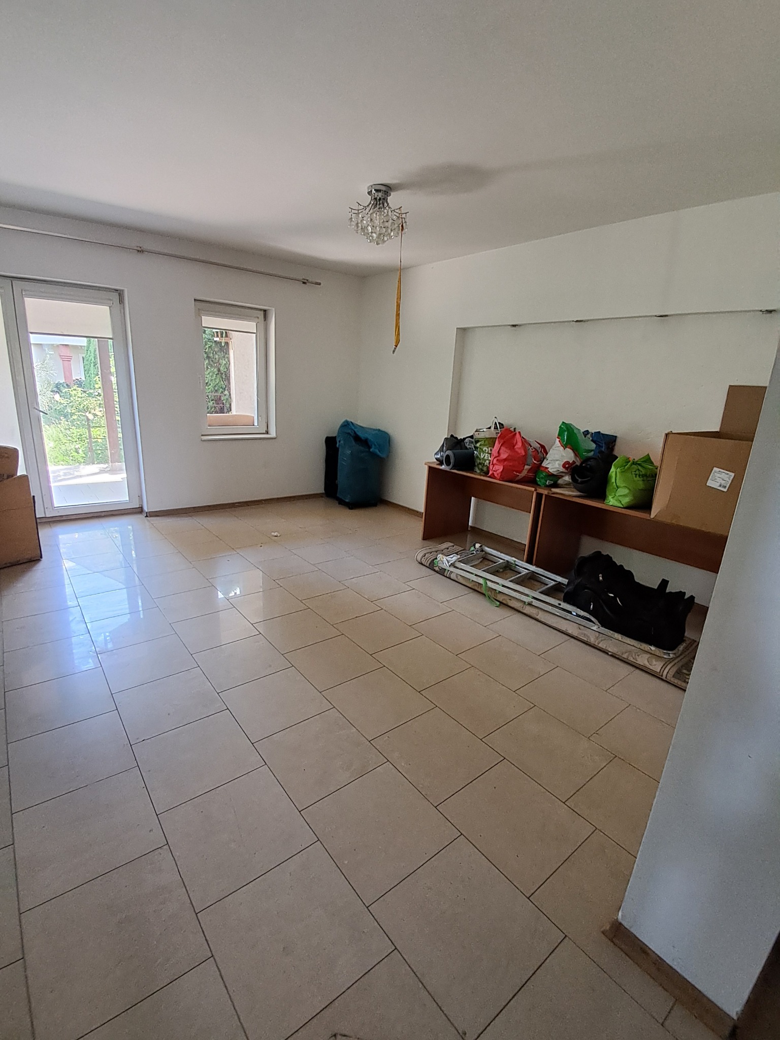 Pusta przestrzeń z beżowymi płytkami podłogowymi, workami na śmieci, kartonami i rozłożoną drabiną opartą o ścianę, przygotowana do posprzątania po remoncie.