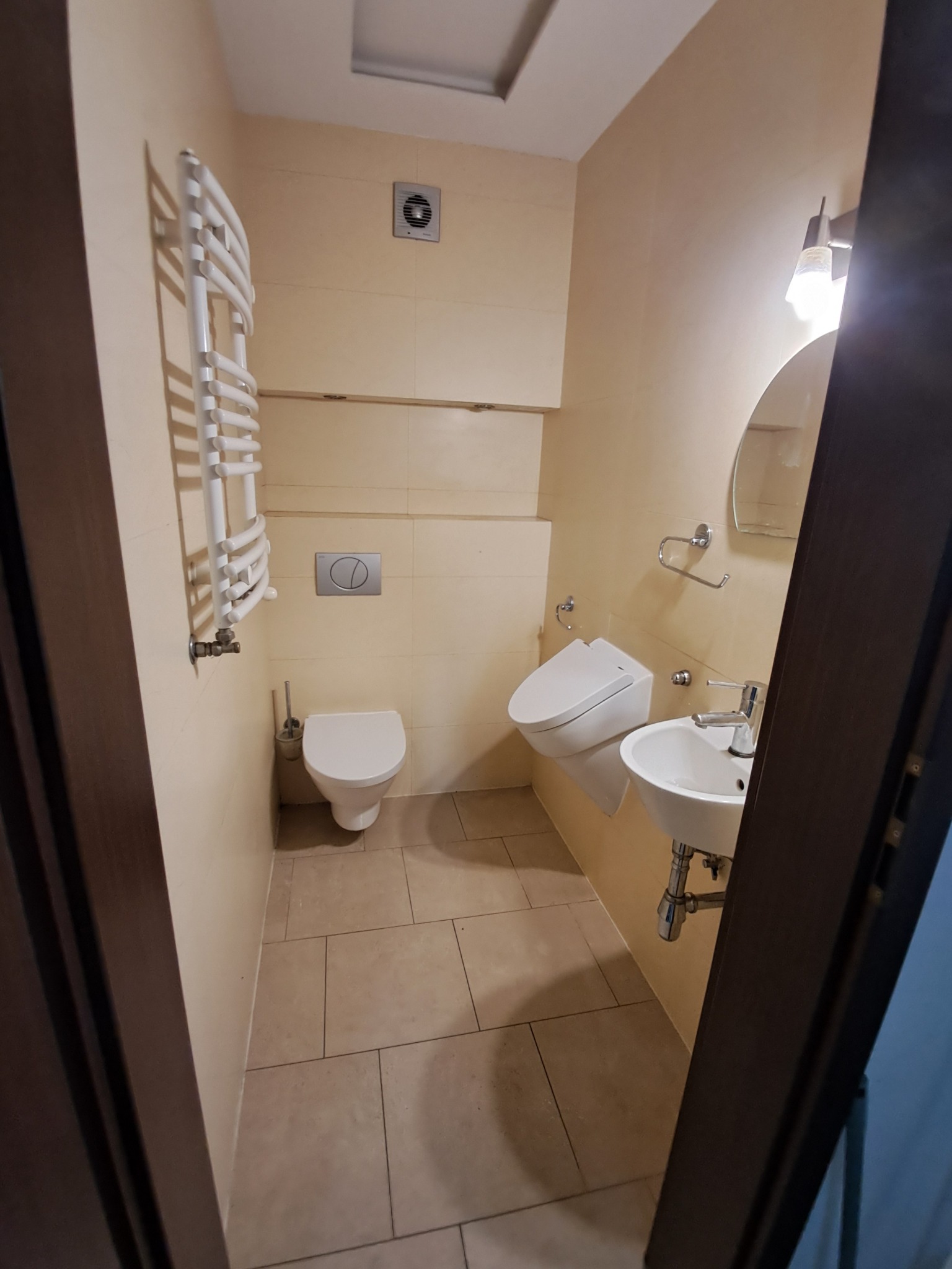 Wnętrze małej łazienki z beżowymi płytkami, widok zza otwartych drzwi na toaletę podwieszaną, umywalkę nablatową i grzejnik drabinkowy.