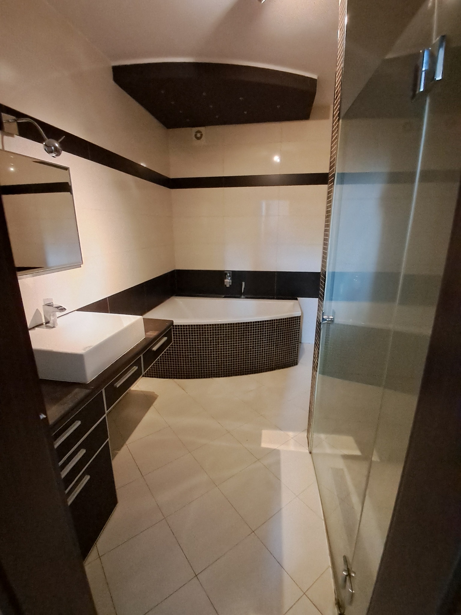 Nowoczesna łazienka z wanną obudowaną mozaiką, białą umywalką nablatową i szklaną kabiną prysznicową, utrzymana w jasnej kolorystyce z czarnymi akcentami.