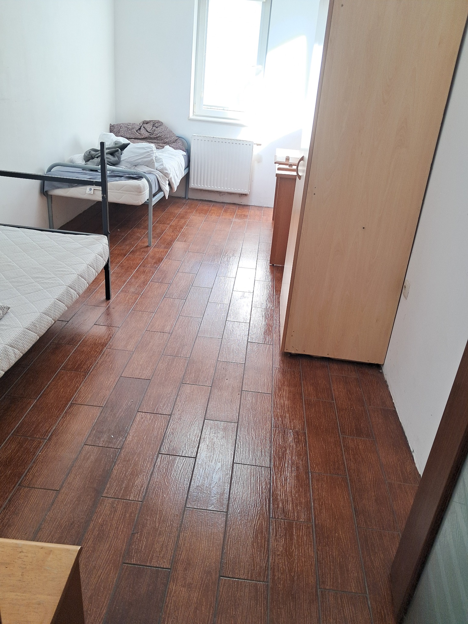 Pokój z dwoma łóżkami i drewnopodobną podłogą, widoczny bałagan na jednym z łóżek, szafa przy ścianie.