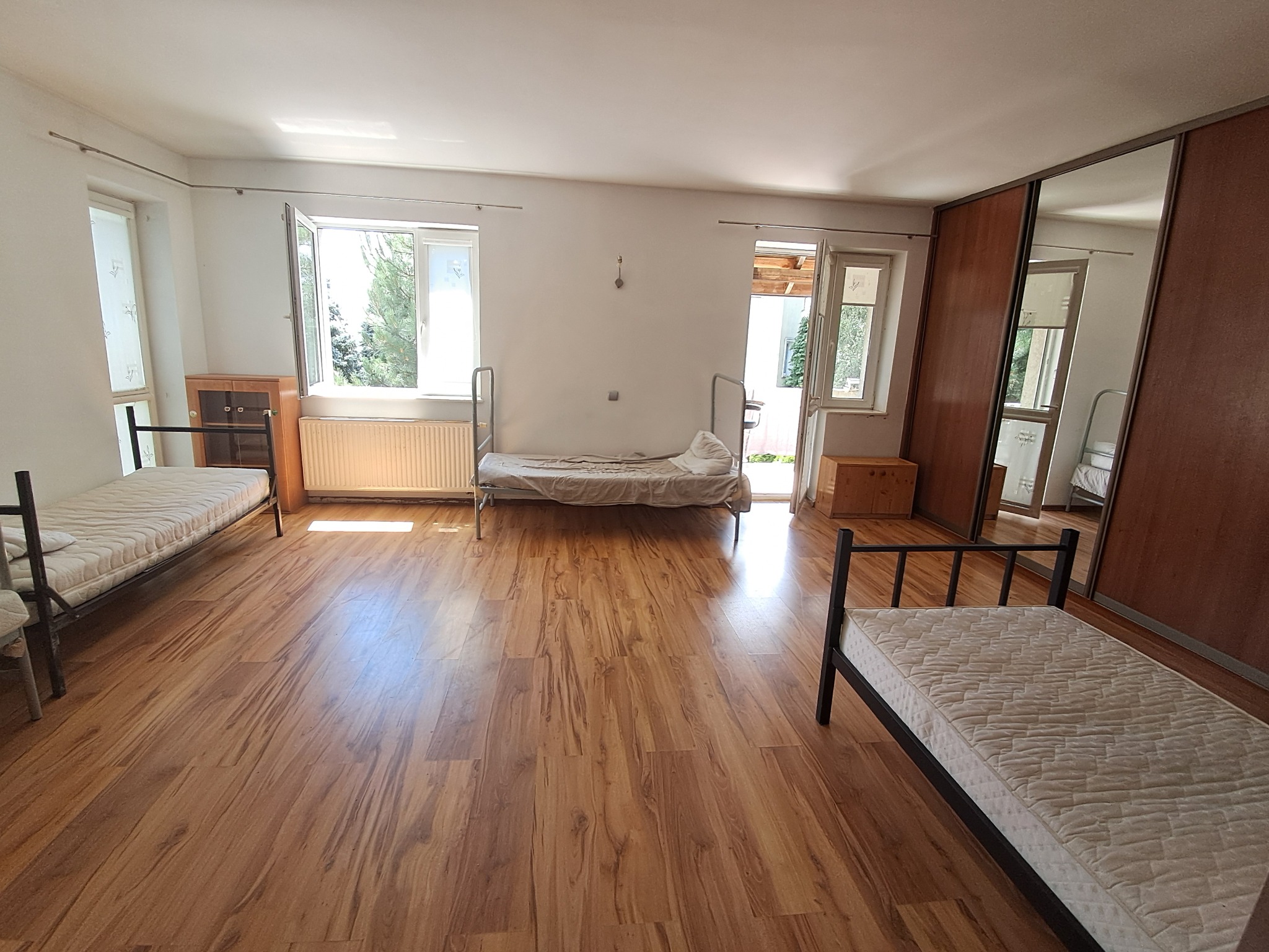 Przestronny pokój z trzema pojedynczymi łóżkami z metalowymi ramami, drewnianą podłogą i szafą z lustrzanymi drzwiami, widok na okno i drzwi balkonowe.