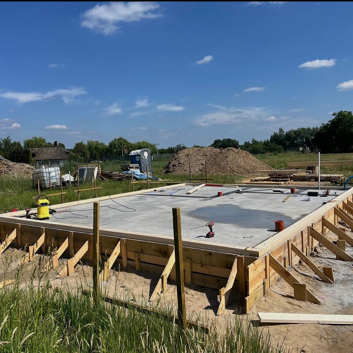 Betonowa płyta fundamentowa z drewnianym szalunkiem pod dom szkieletowy, widoczna wiertarka akumulatorowa i poziomica laserowa, w tle przenośna toaleta i pryzma ziemi.