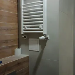 Biały, drabinkowy grzejnik łazienkowy zamontowany na szarej ścianie z płytek, obok drewnopodobnej zabudowy z widocznym przyciskiem spłuczki i rolką papieru toaletowego.