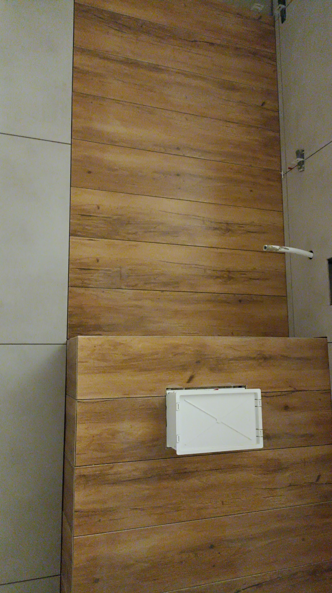Ściana łazienki wyłożona płytkami imitującymi drewno w ciepłych odcieniach brązu, z widocznym białym otworem rewizyjnym i fragmentem szarej ściany z płytkami w tle.