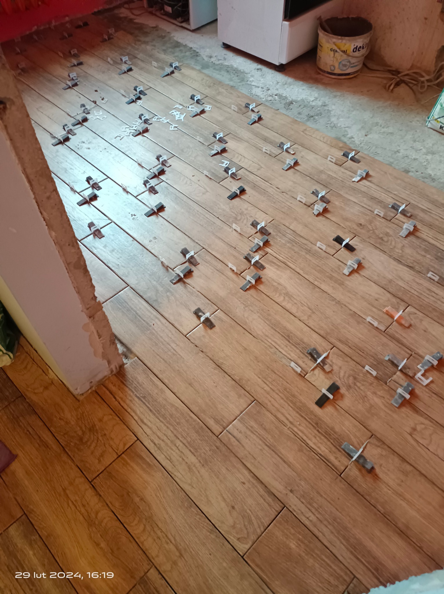 Układanie płytek imitujących drewno na podłodze, widoczne krzyżyki dystansowe i fragment ściany w trakcie prac wykończeniowych.