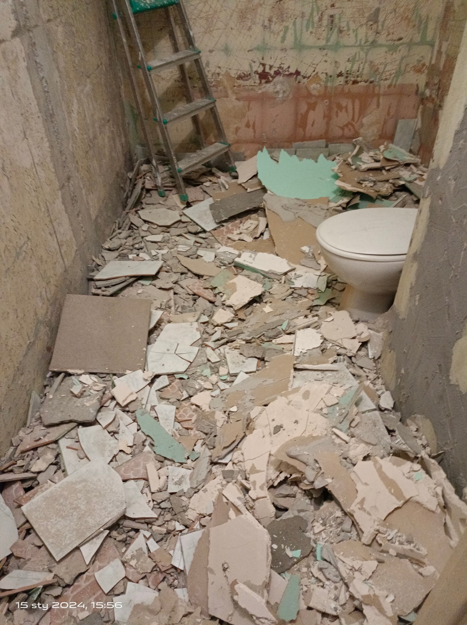 Łazienka w trakcie demontażu: gruz po starych płytkach, fragment toalety i drabina oparta o ścianę z resztkami farby.