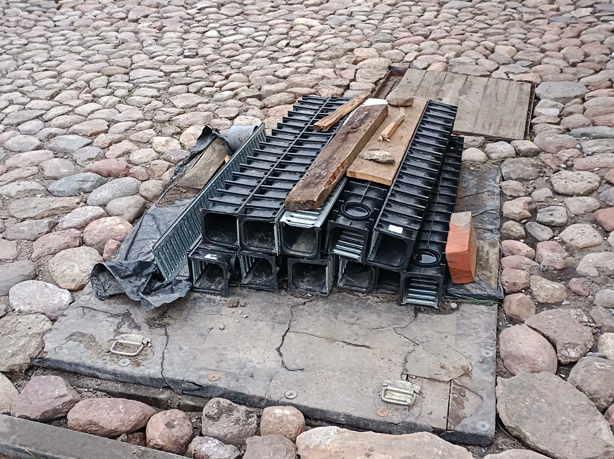 Stos czarnych, plastikowych korytek kablowych ułożonych na betonowej płycie z metalowymi uchwytami, obok cegła i drewniane deski, otoczone brukowaną nawierzchnią z kamieni polnych.