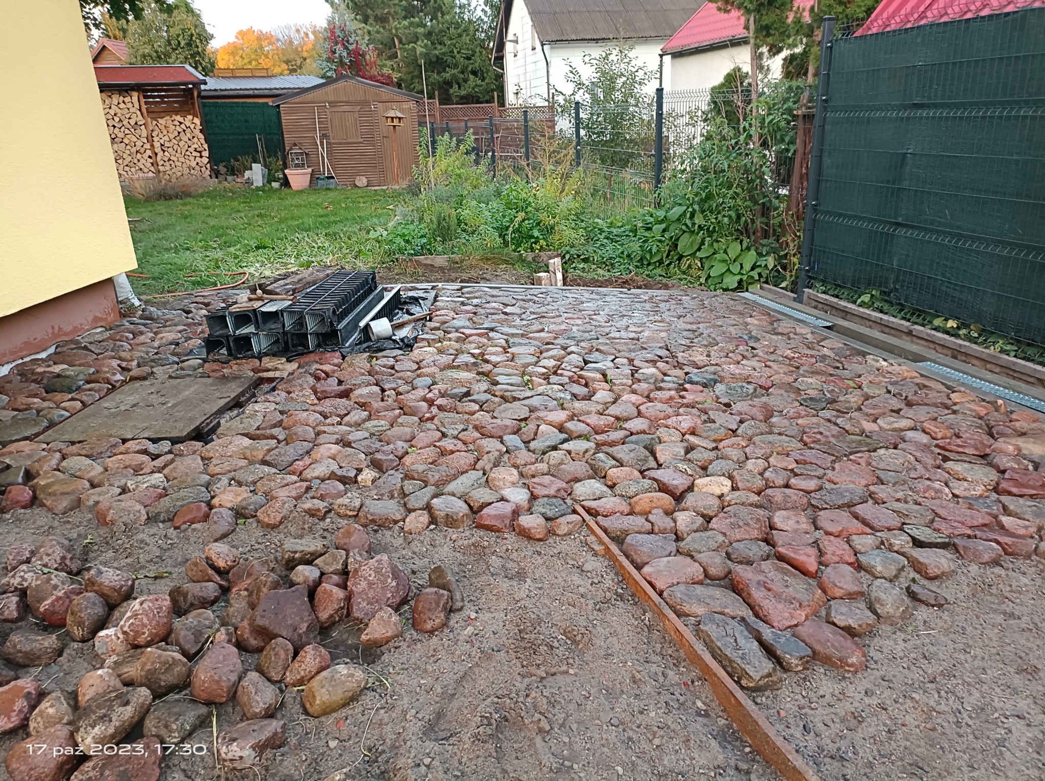 Układanie nawierzchni z kamieni polnych na zewnątrz budynku, widoczne metalowe profile i stos metalowych elementów w tle ogrodu.