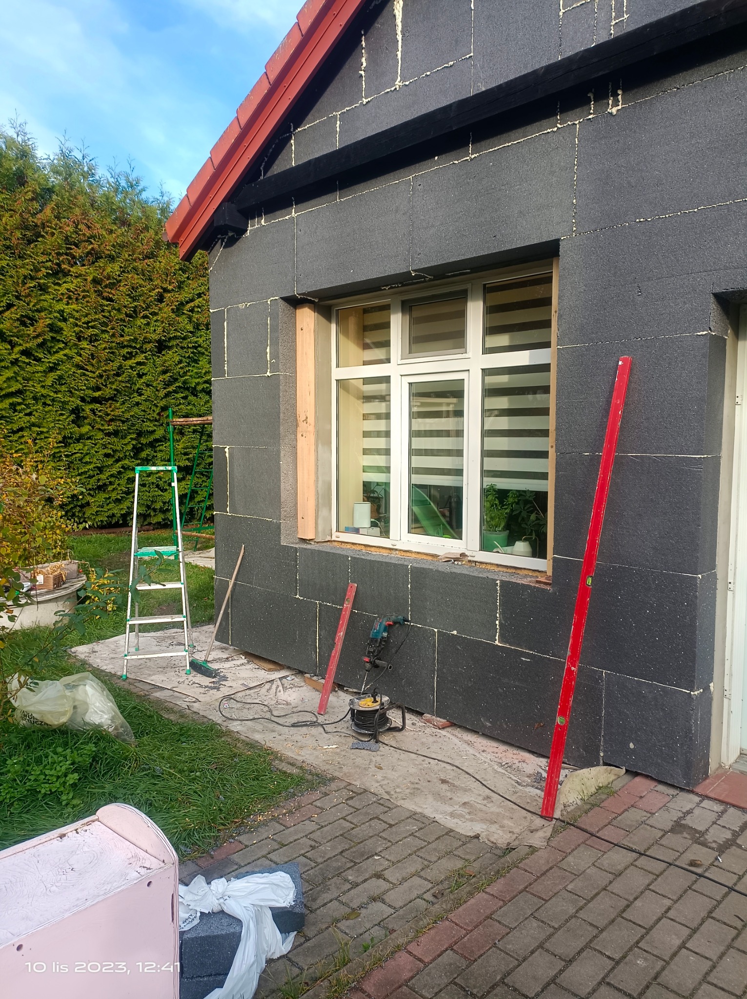 Dom w trakcie ocieplania, ściany pokryte czarnym styropianem, widoczne narzędzia: poziomica, wiertarka, drabina, szczotka. Okno z żaluzjami częściowo osadzone w drewnianej ramie.