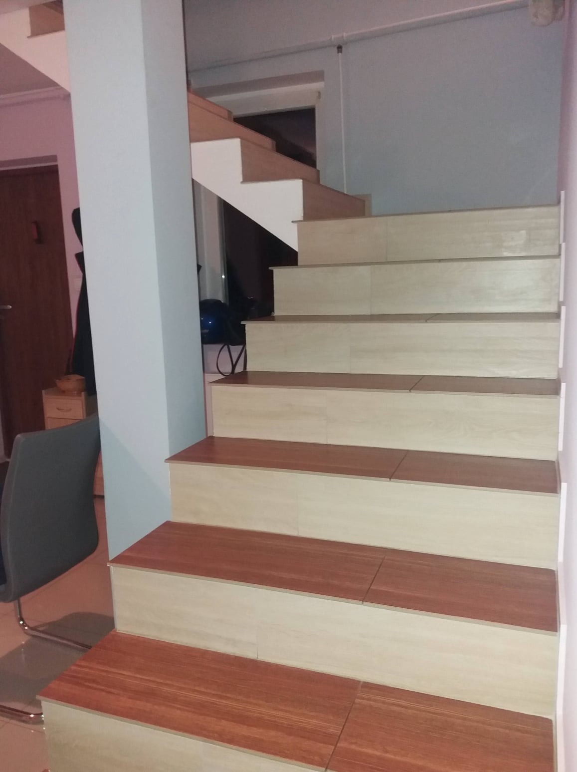 Schody wewnętrzne z drewnopodobnymi płytkami, widoczne stopnie w dwóch odcieniach brązu i beżu, ułożone w poprzek, z kolumną wspierającą konstrukcję.