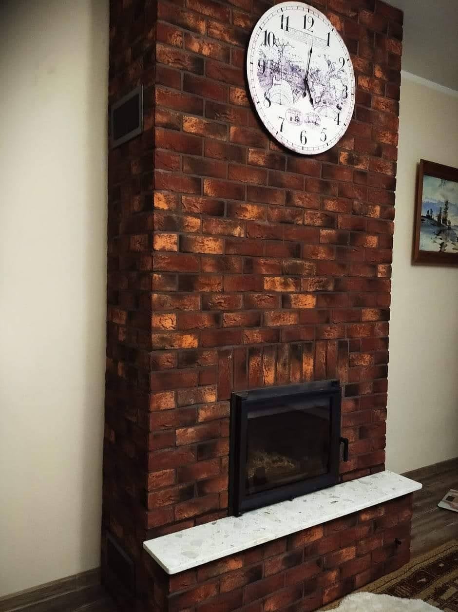 Kominek obudowany czerwoną cegłą klinkierową z czarnym wkładem kominkowym i blatem z białego marmuru, nad nim zegar ścienny z mapą świata.