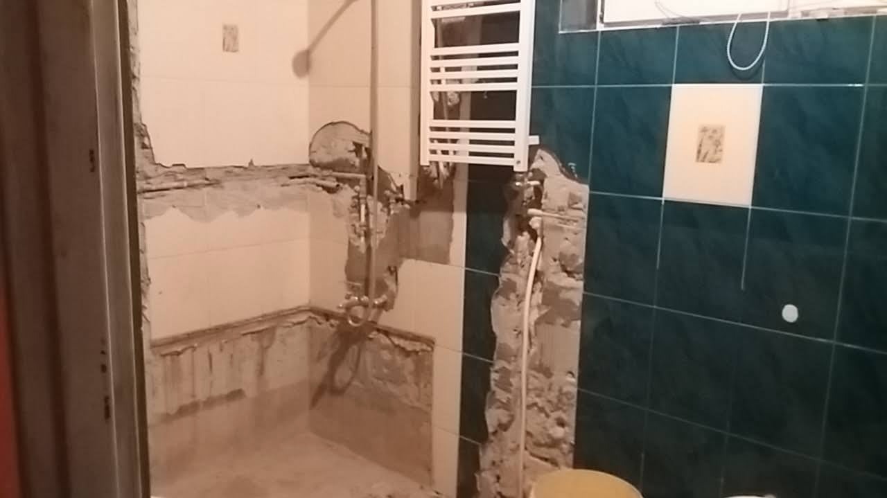 Demontaż płytek w łazience, widoczne fragmenty instalacji wodnej i grzejnik drabinkowy na ścianie pokrytej zielonymi kafelkami z białym akcentem.