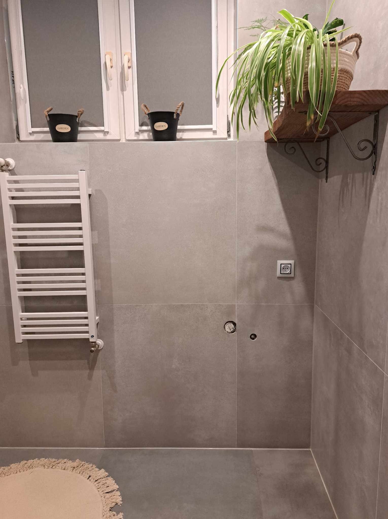 Wykończona łazienka z szarymi płytkami, grzejnikiem drabinkowym, drewnianą półką z rośliną doniczkową w plecionym koszu oraz detalami dekoracyjnymi w postaci dwóch czarnych doniczek na parapecie.