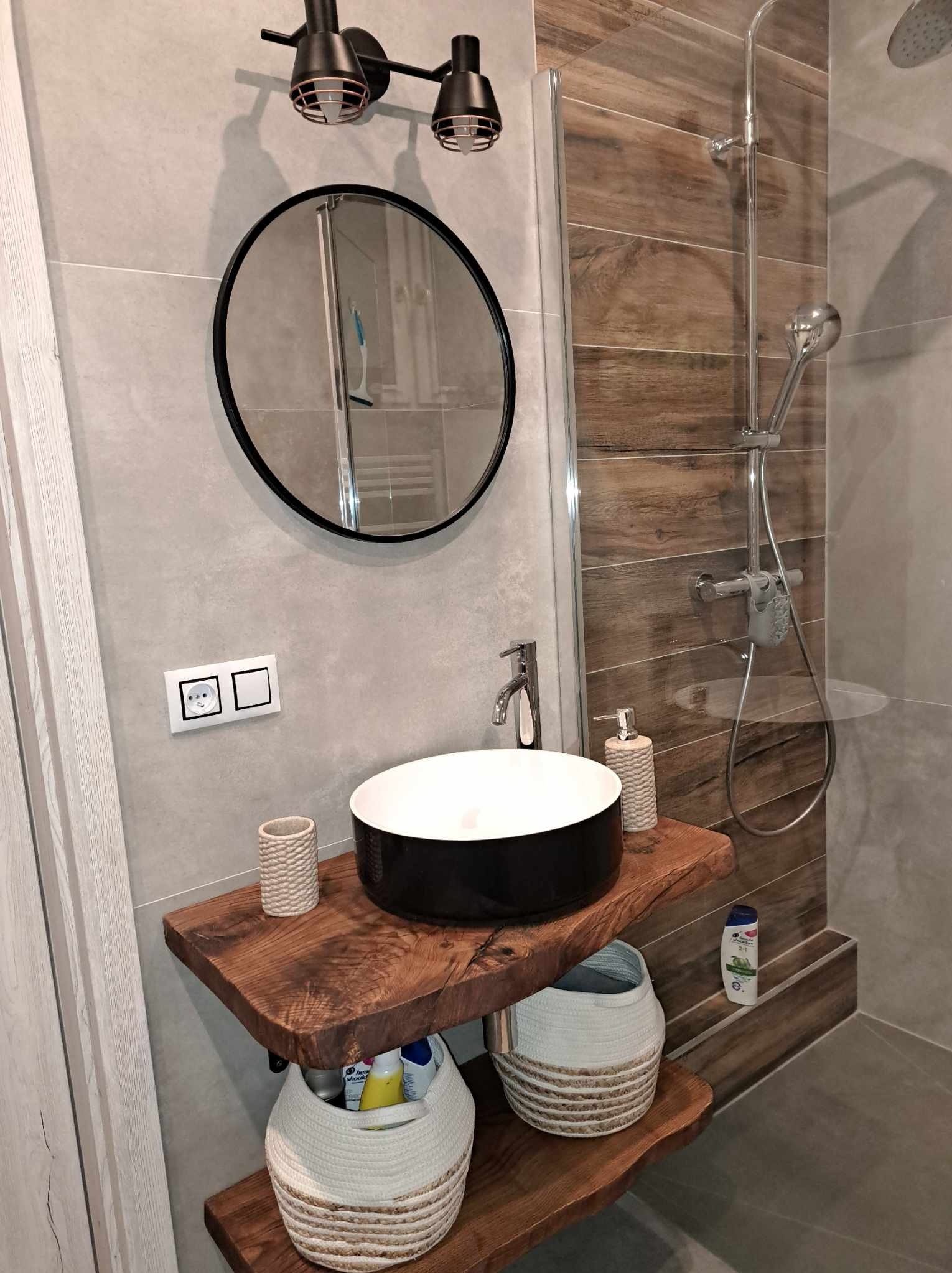 Nowoczesna łazienka z okrągłym lustrem w czarnej ramie, umywalką nablatową na drewnianym blacie z półkami, oraz kabiną prysznicową z panelami imitującymi drewno.