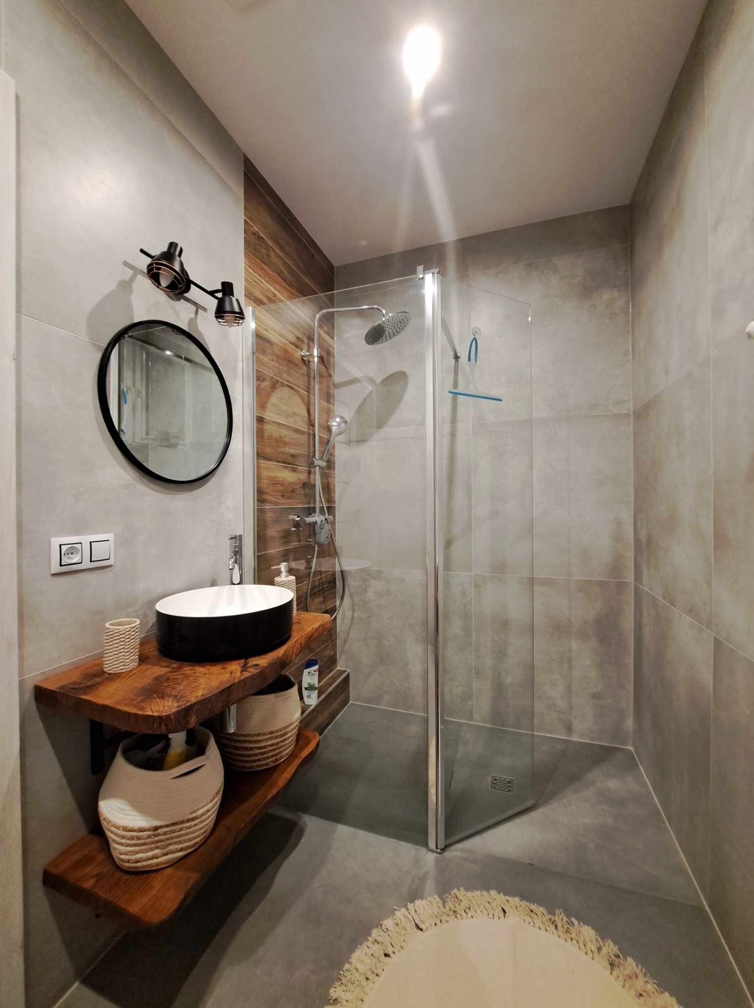 Nowoczesna łazienka z kabiną prysznicową, umywalką nablatową na drewnianym blacie i okrągłym lustrem, oświetlona czarnymi lampami punktowymi, z szarymi płytkami i drewnianą ścianą w strefie prysznica.