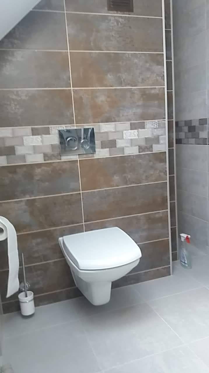 Wisząca toaleta w nowoczesnej łazience z brązowo-szarymi płytkami, dekoracyjnym pasem mozaiki i chromowanym przyciskiem spłukiwania.