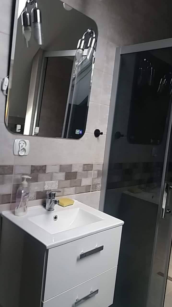 Nowoczesna łazienka z białą szafką pod umywalką, lustrem z oświetleniem LED i kabiną prysznicową z ciemnego szkła, wykończona płytkami w odcieniach szarości.