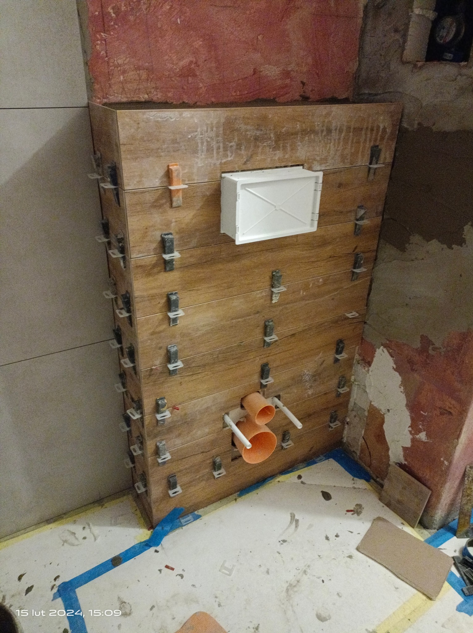 Obrudowana konstrukcja z płytek imitujących drewno, z widocznymi rurami instalacyjnymi i białą klapą rewizyjną, zamocowana przy użyciu klipsów poziomujących, w trakcie prac wykończeniowych w łazience.