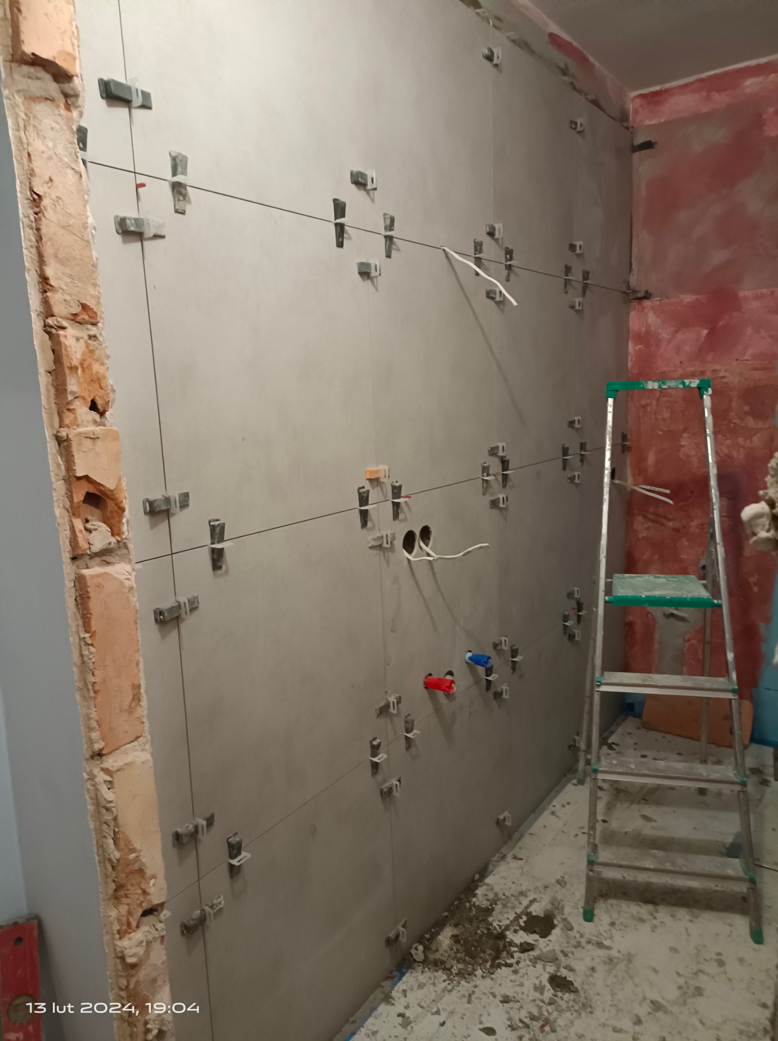 Montaż płytek na ścianie w łazience, widoczne krzyżyki dystansowe, poziomowanie sznurkiem, surowy mur z cegły obok gładkiej ściany, przygotowane otwory na instalację wodną z czerwonym i niebieskim...