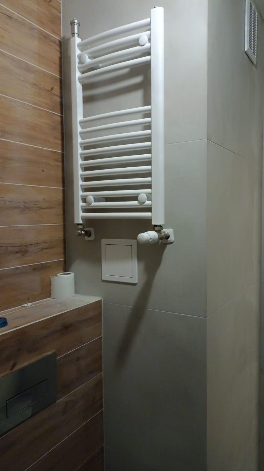 Biały, drabinkowy grzejnik łazienkowy zamontowany na szarej ścianie z płytek, obok drewnopodobnej zabudowy z widocznym przyciskiem spłuczki i rolką papieru toaletowego.