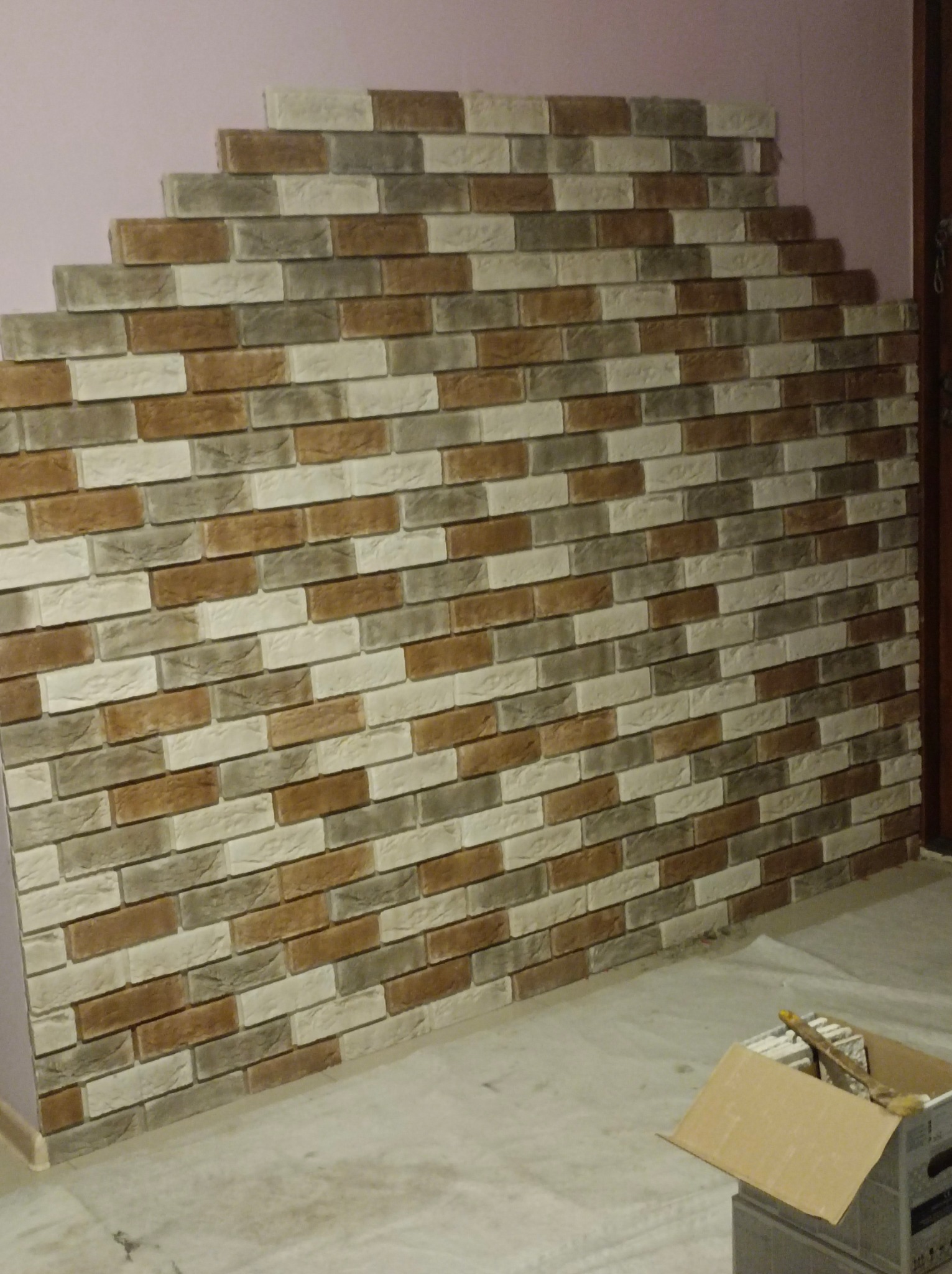 Ściana pokryta dekoracyjną okładziną z cegieł w odcieniach brązu, szarości i beżu, układana schodkowo na tle fioletowej ściany, z kartonem płytek na podłodze.