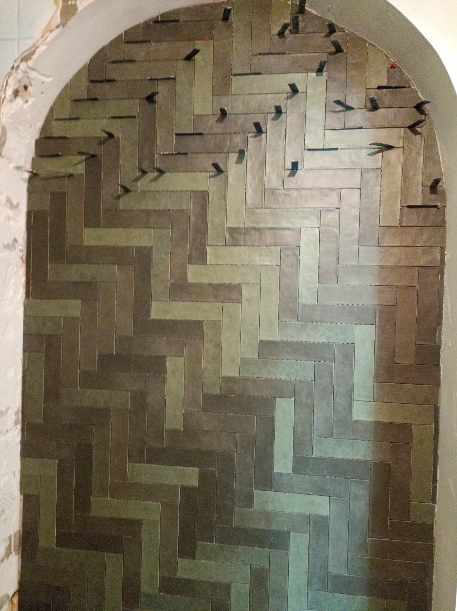 Układanie płytek w jodełkę w niszy łazienkowej, z widocznymi klipsami poziomującymi, na nierównym tle surowej ściany.